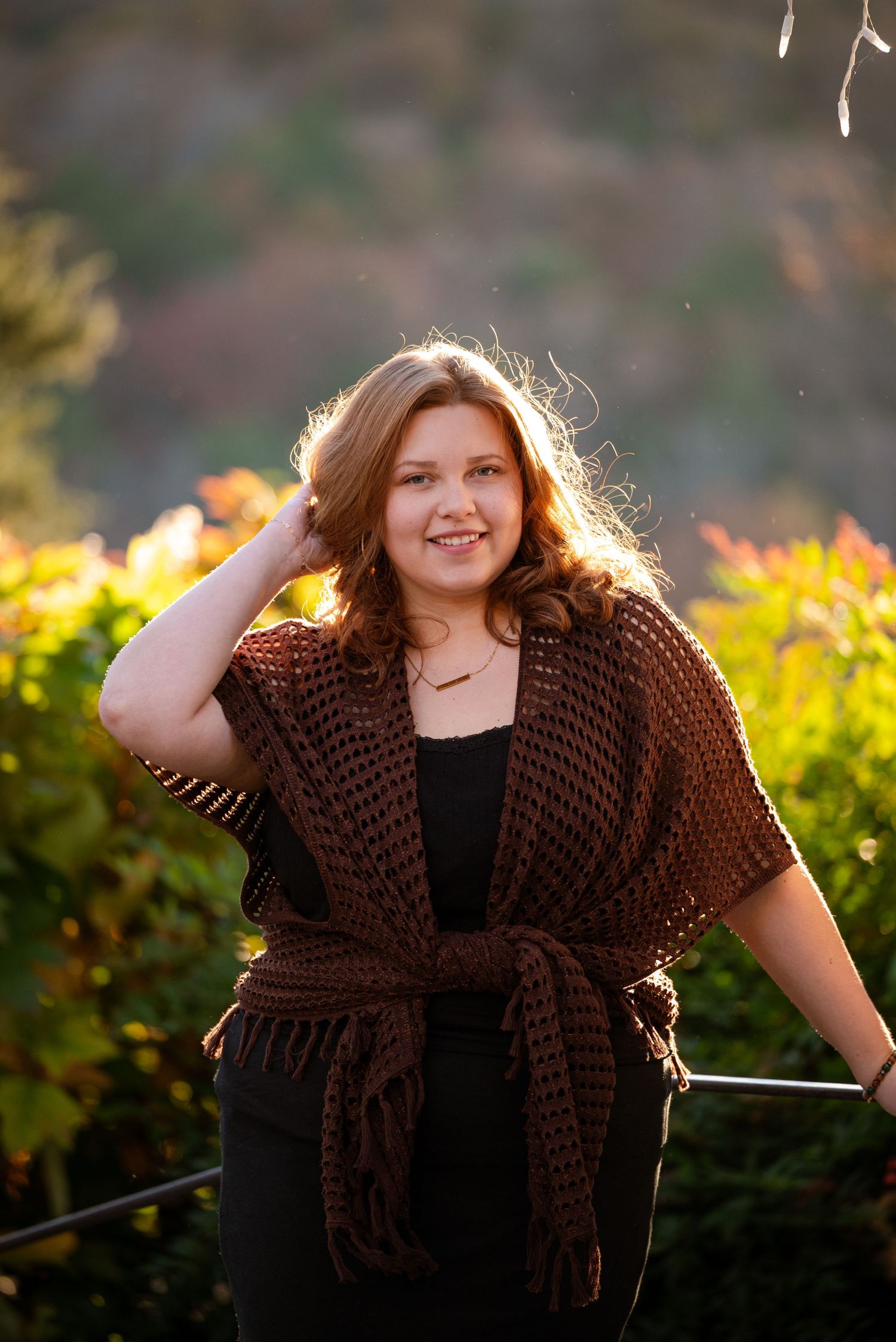 Young woman in brown knit wrap smiling outdoors with golden autumn bokeh background.