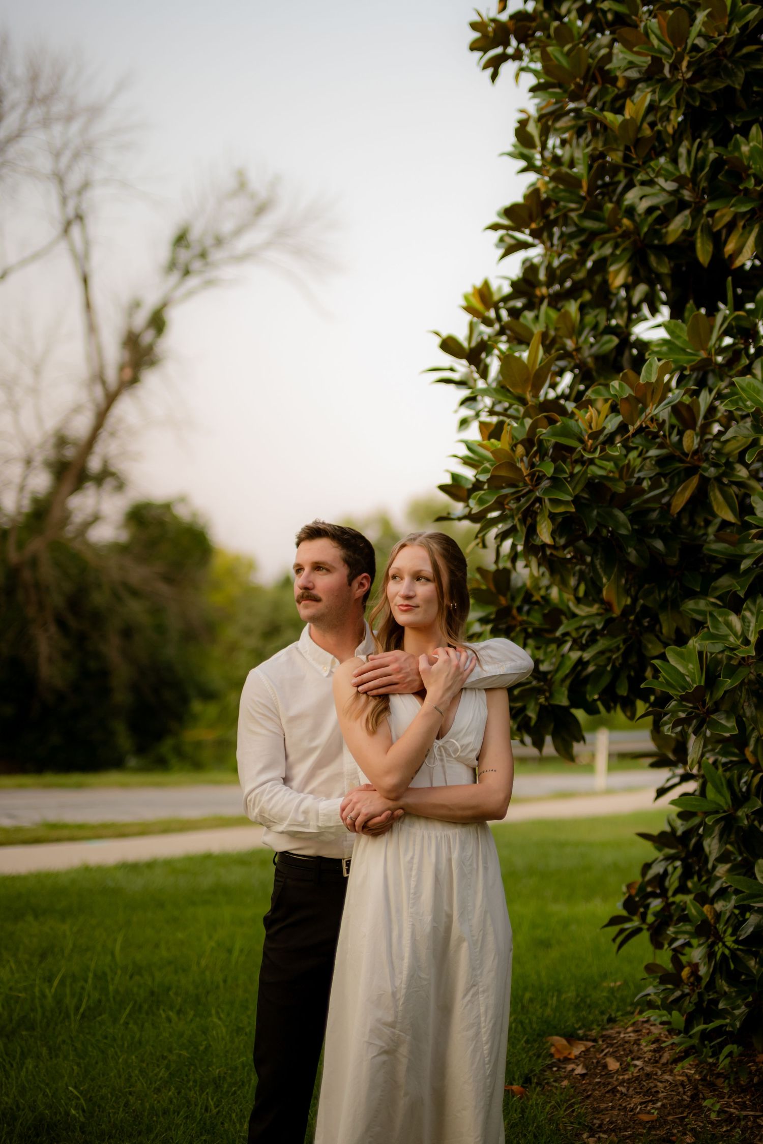 A couple embraces near a magnolia tree in a lush green park, the woman in a white dress gazing dreamily into the distance.