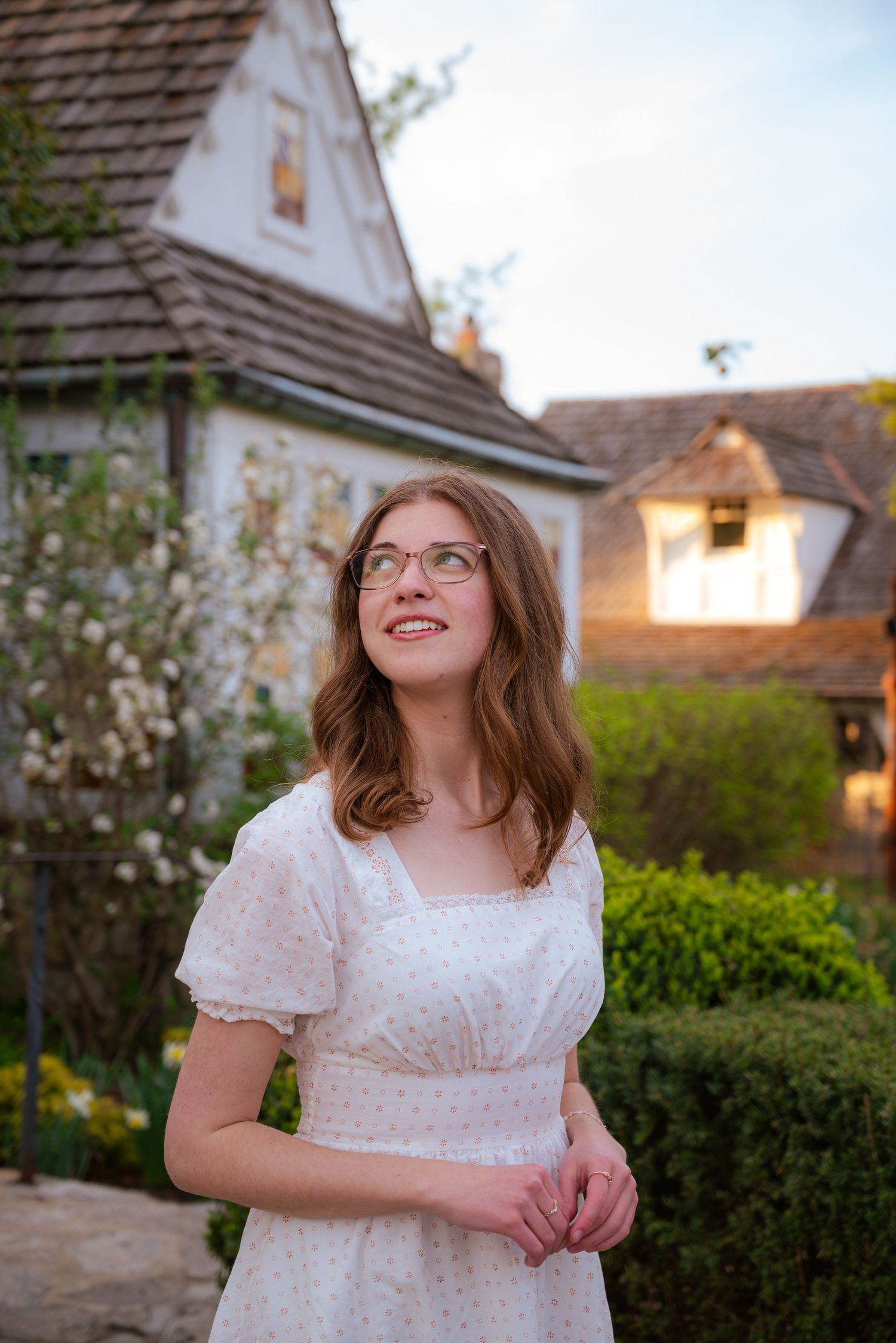 Young woman in white floral dress with glasses smiles outdoors near a charming cottage garden.
