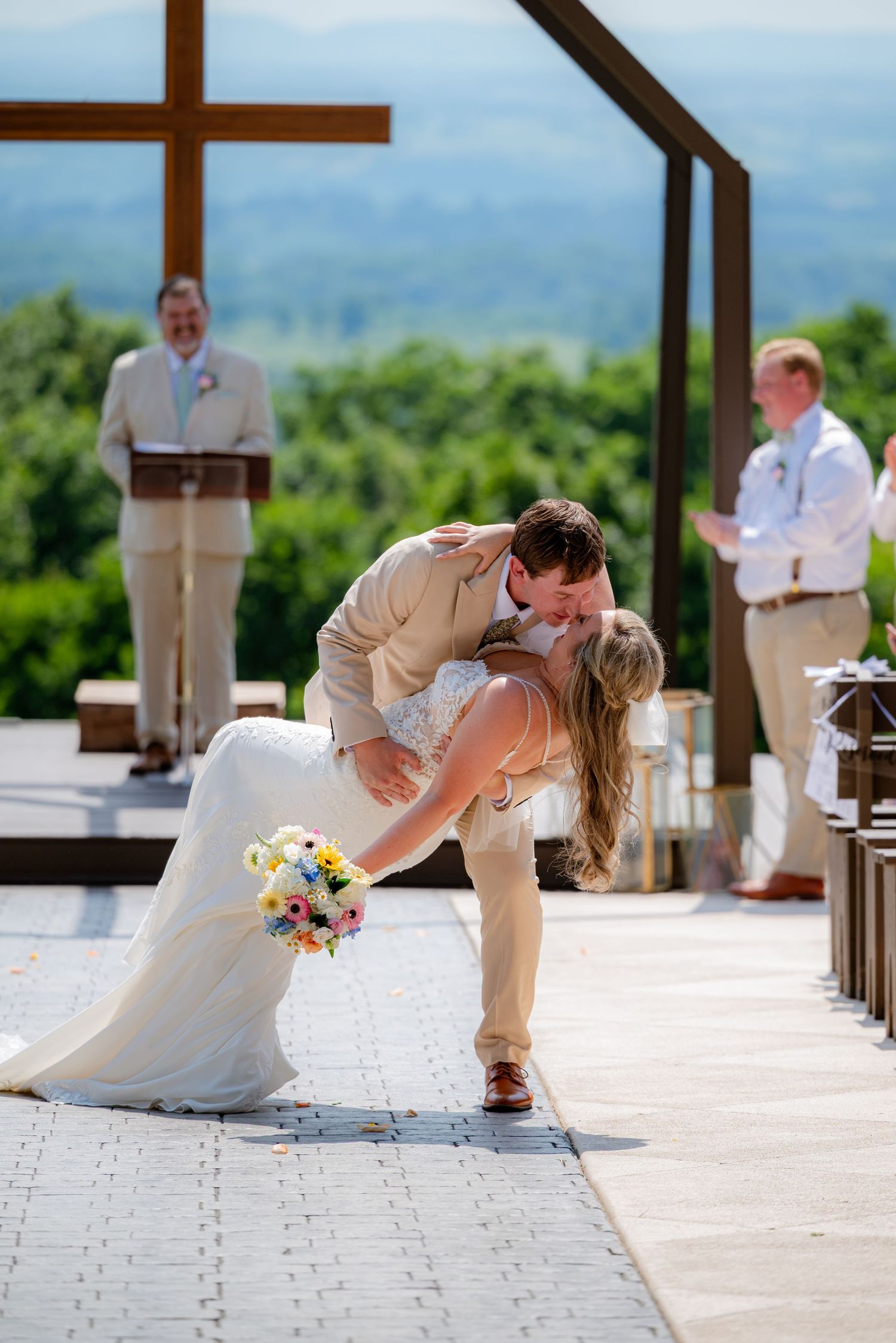 Bride and groom share a romantic dip kiss at outdoor wedding ceremony with mountain views and large cross backdrop.