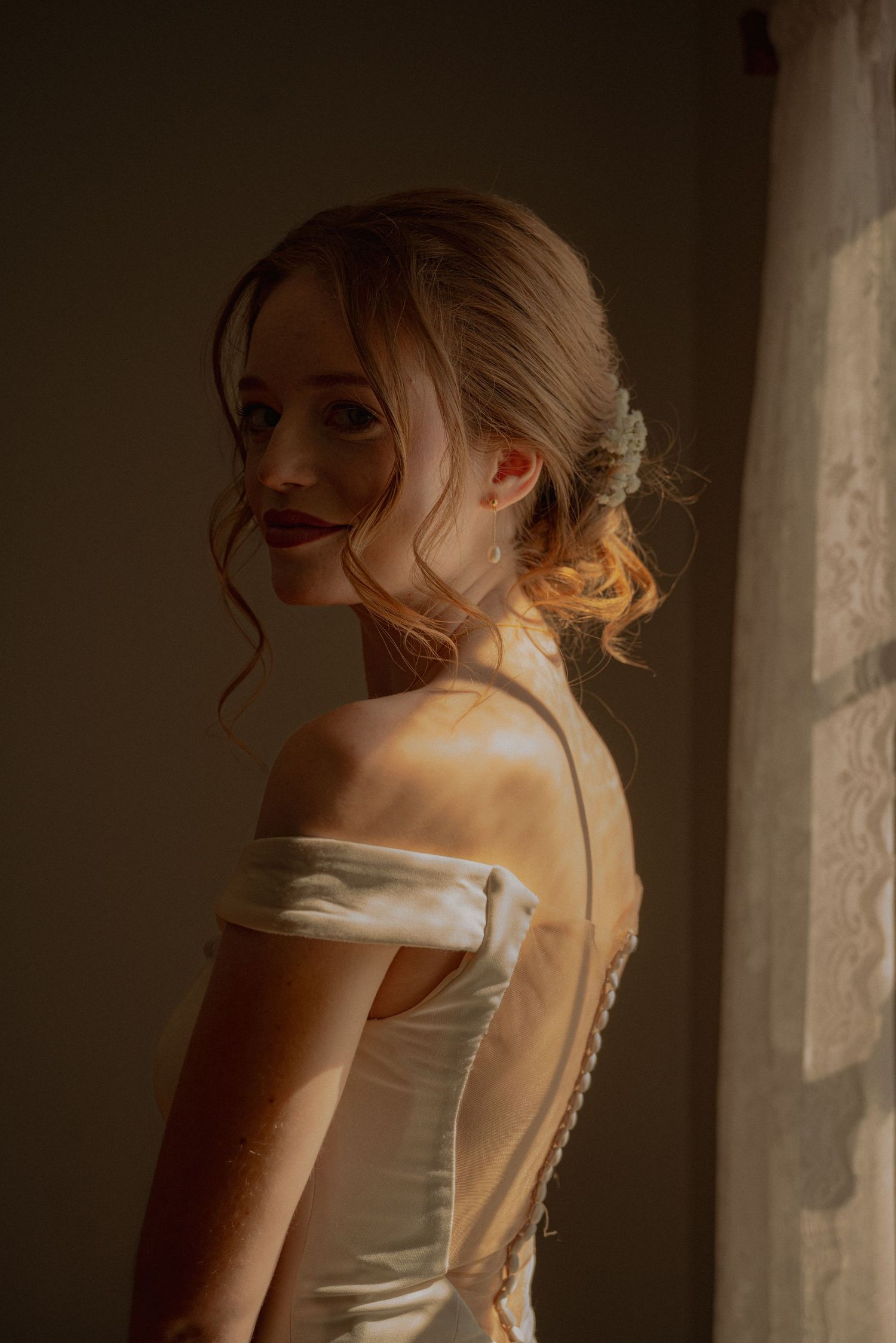 Bride in off-shoulder satin gown with updo hairstyle poses gracefully by a window in soft golden light.