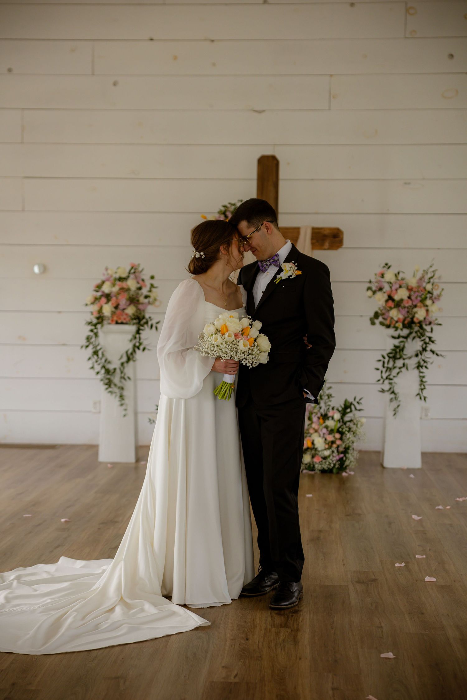 Bride and groom share a romantic kiss at the altar, surrounded by floral arrangements and a wooden cross in a white barn venue.
