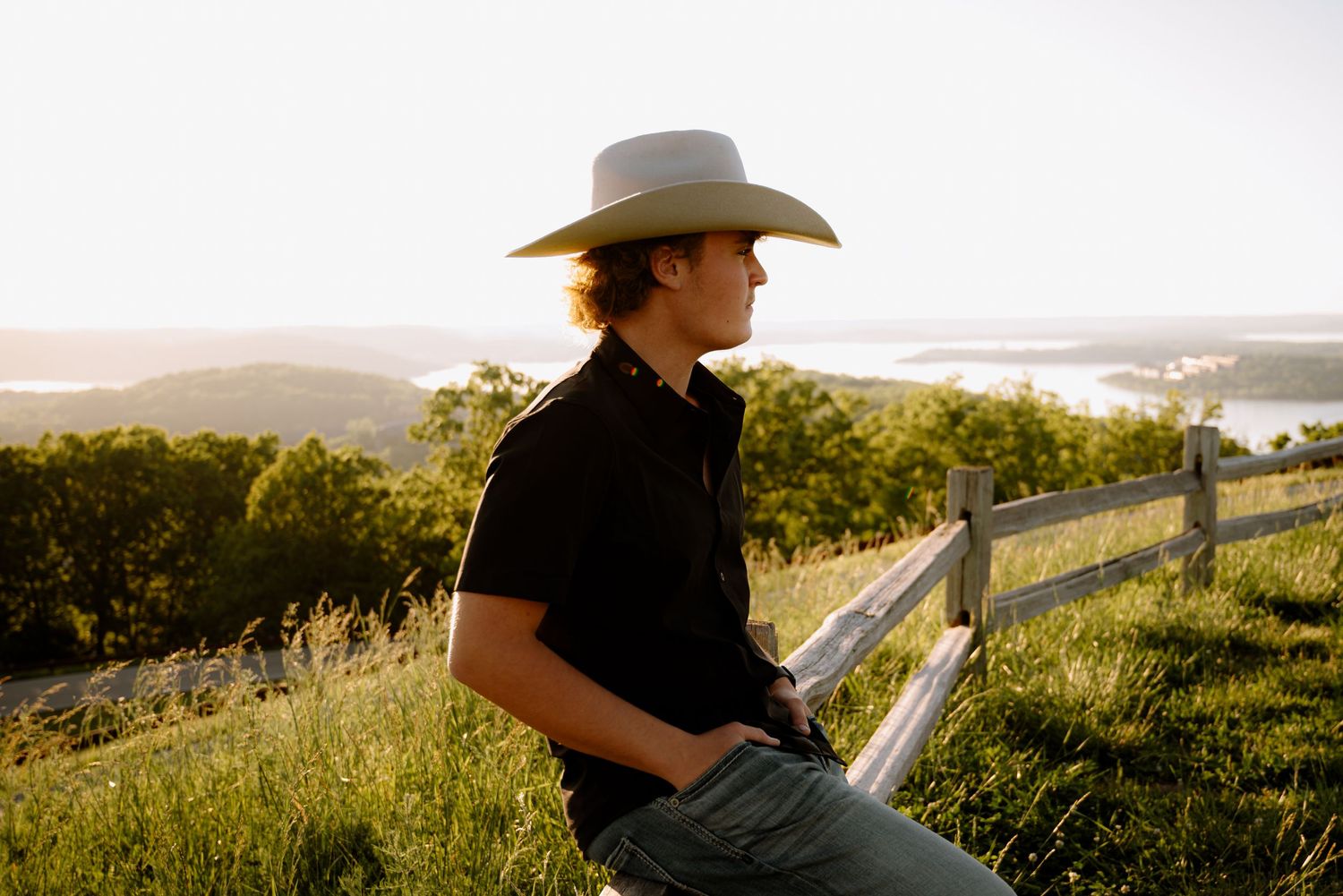 A young man in a white cowboy hat and black shirt sits on a wooden fence overlooking a scenic valley.