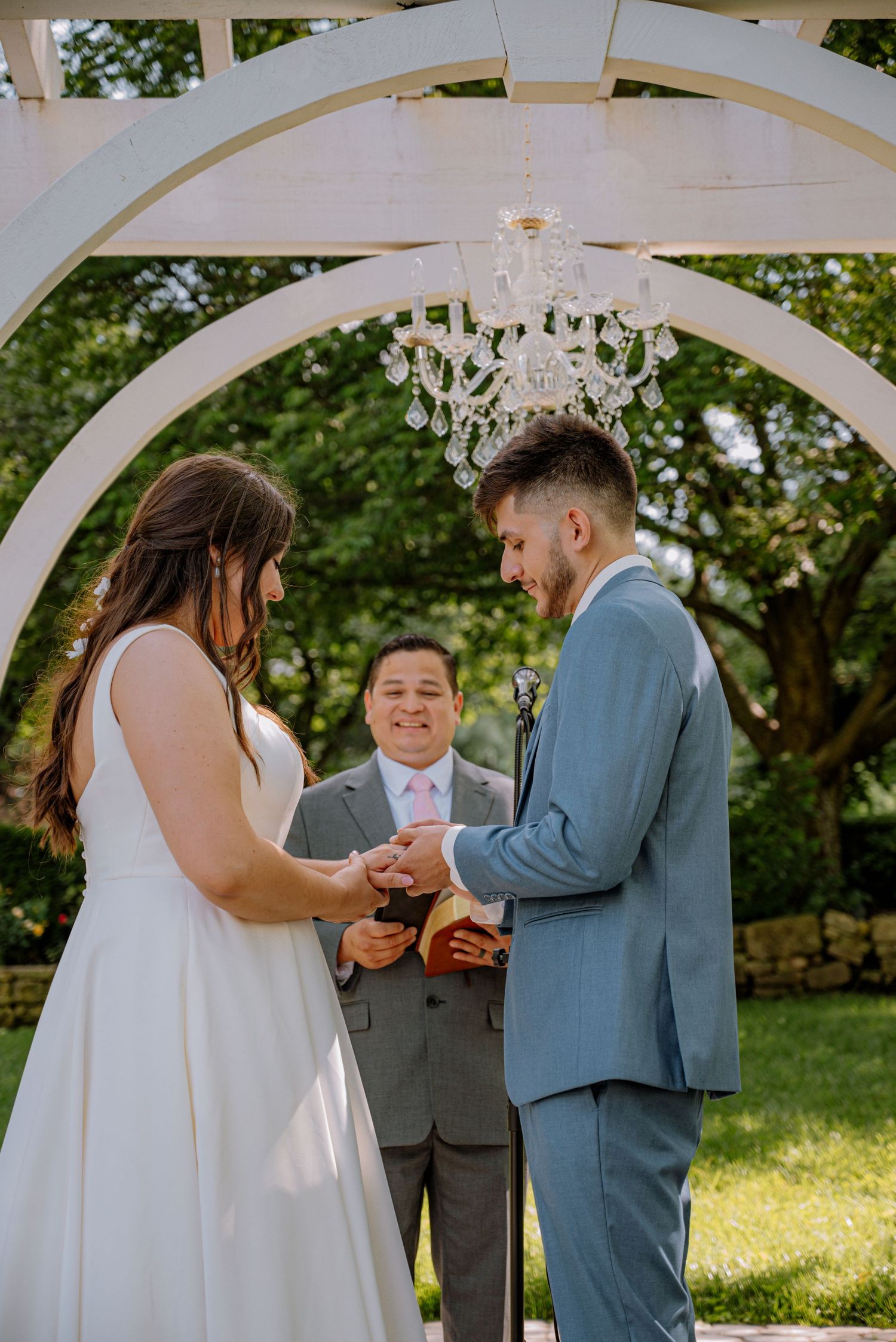 Bride and groom exchanging rings during their outdoor ceremony with a beautiful chandelier above them.