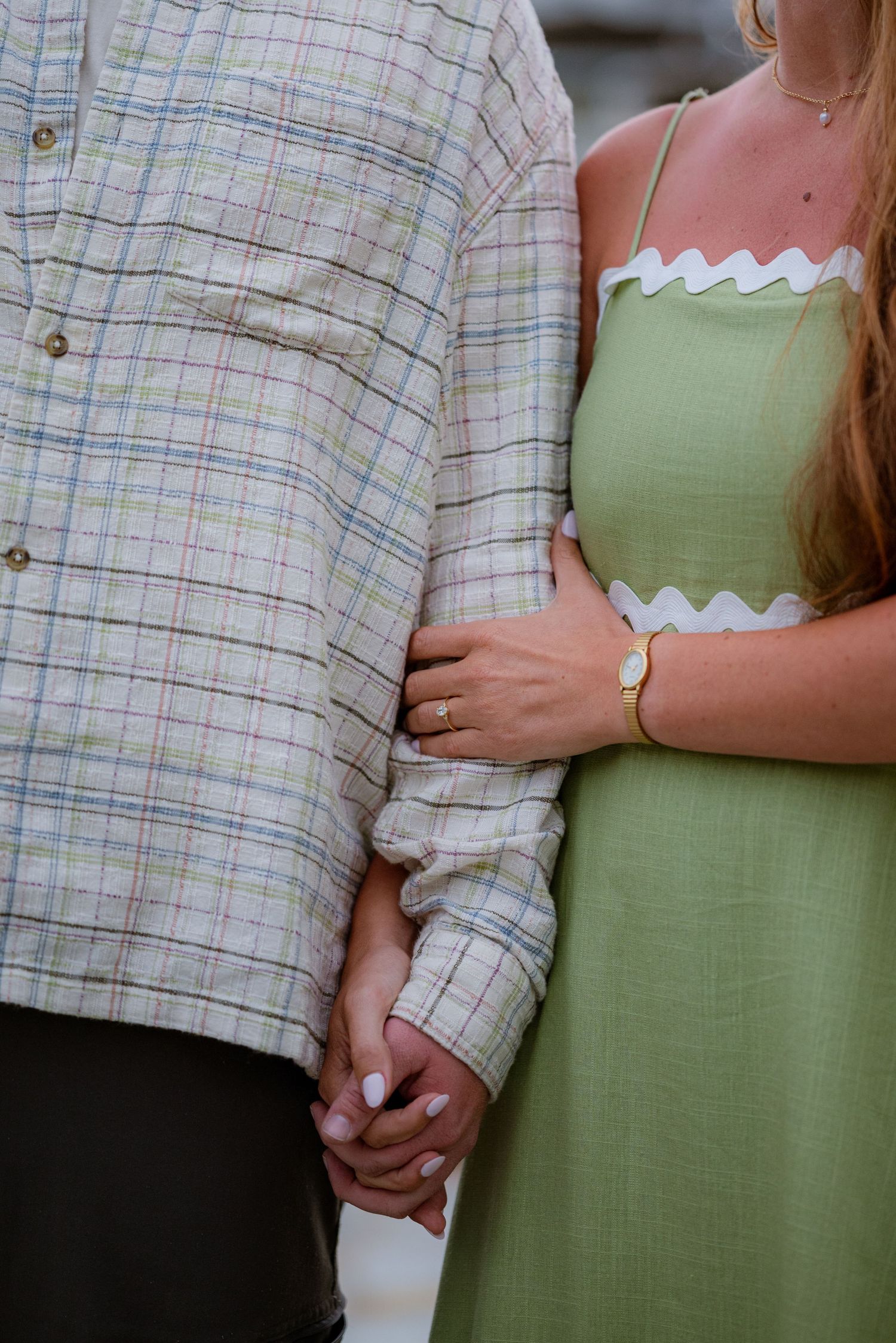 Couple holding hands, woman in green zig-zag trim dress with gold watch, man in plaid shirt, romantic outdoor photo.