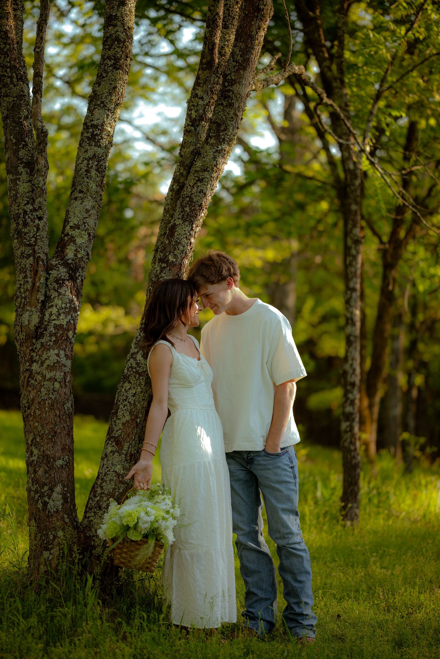 Couple in white dress and jeans share a romantic moment leaning against a tree in a lush green forest.