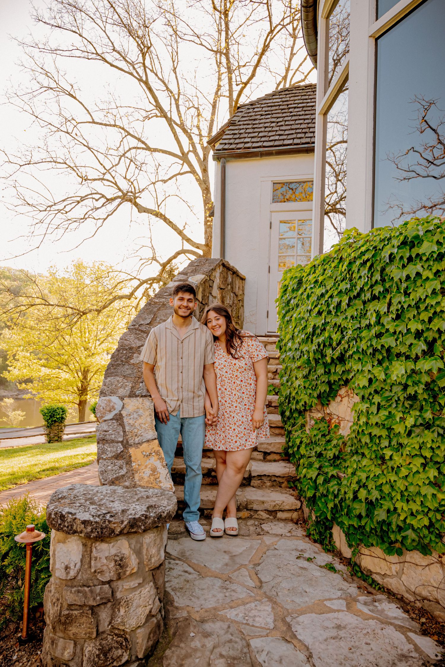 Smiling engaged couple standing on stone steps outside a charming ivy-covered home at sunset.