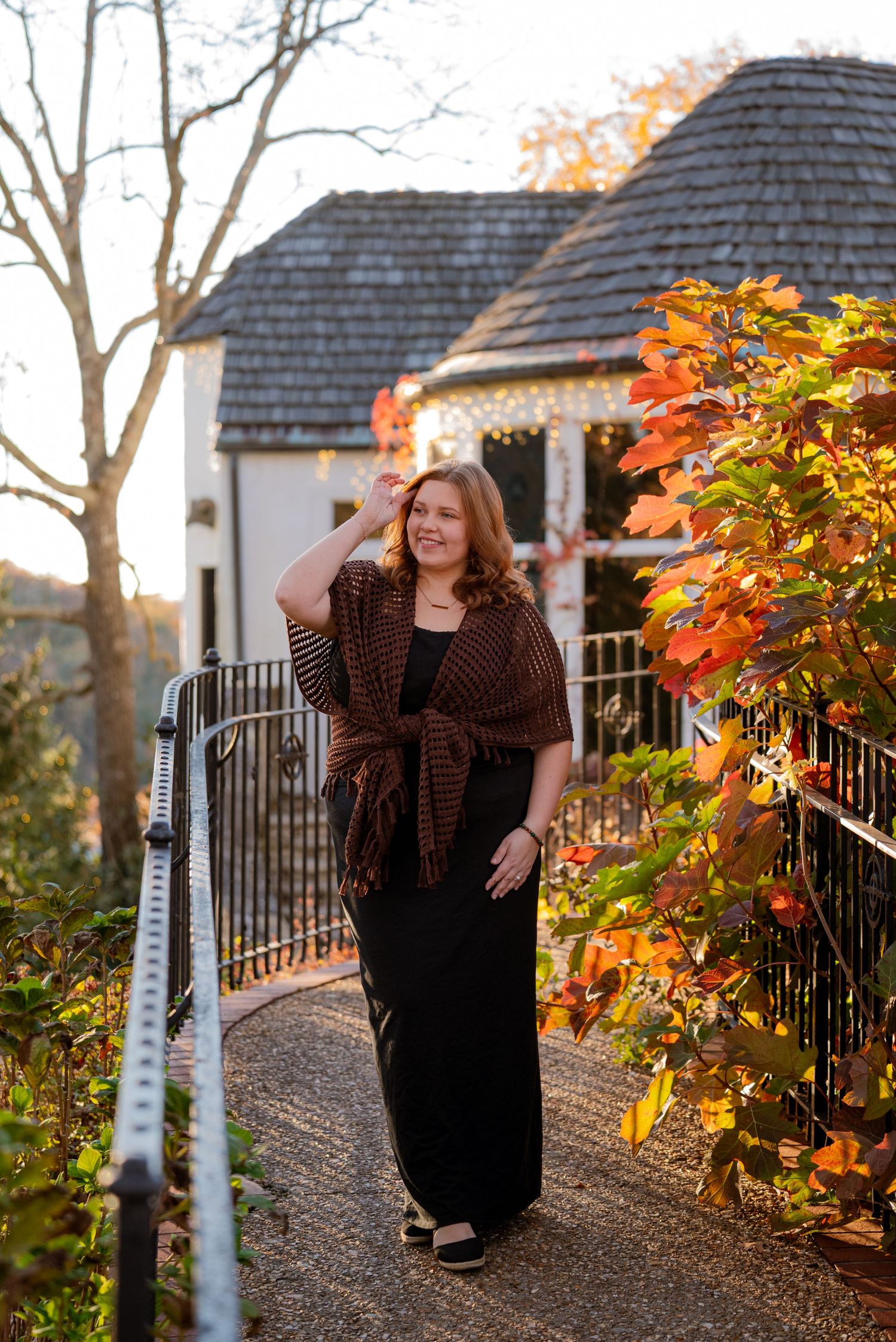 Woman in black dress and brown fringe shawl walking path near gazebo with fall foliage.