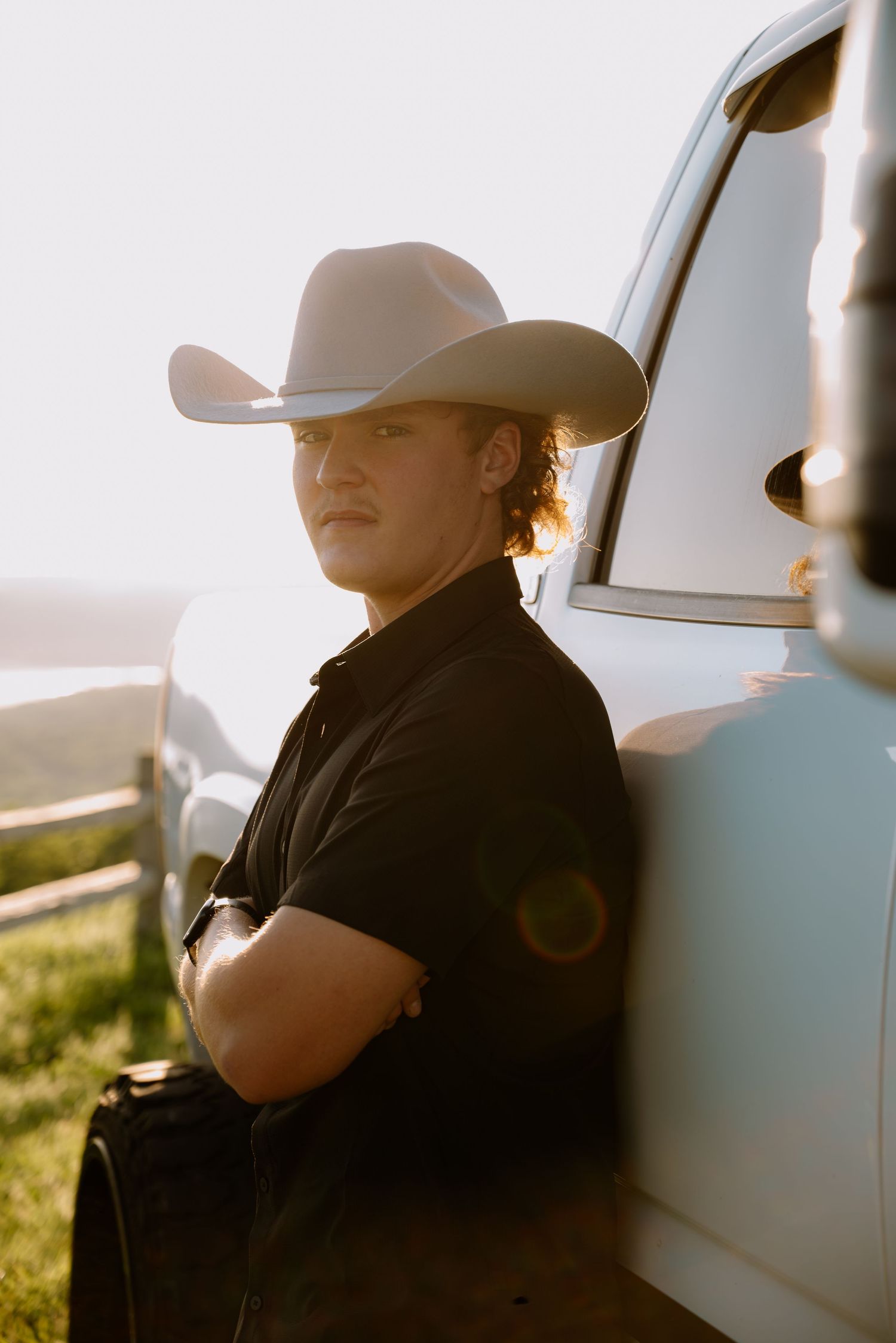 A young man in a black shirt and white cowboy hat leans against a white truck at sunset.