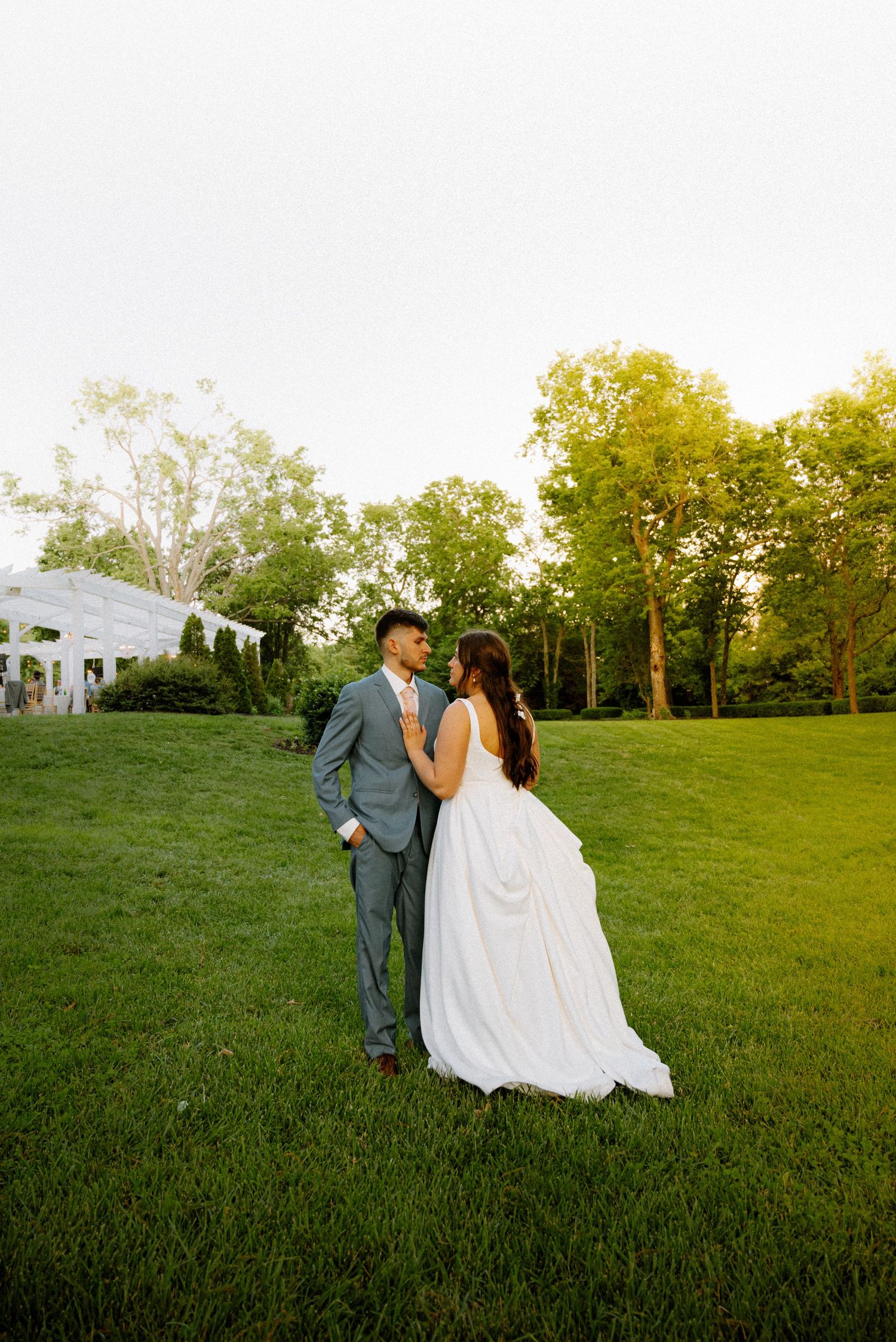 Bride and groom share a romantic moment on lush green lawn at golden hour outdoor wedding venue.