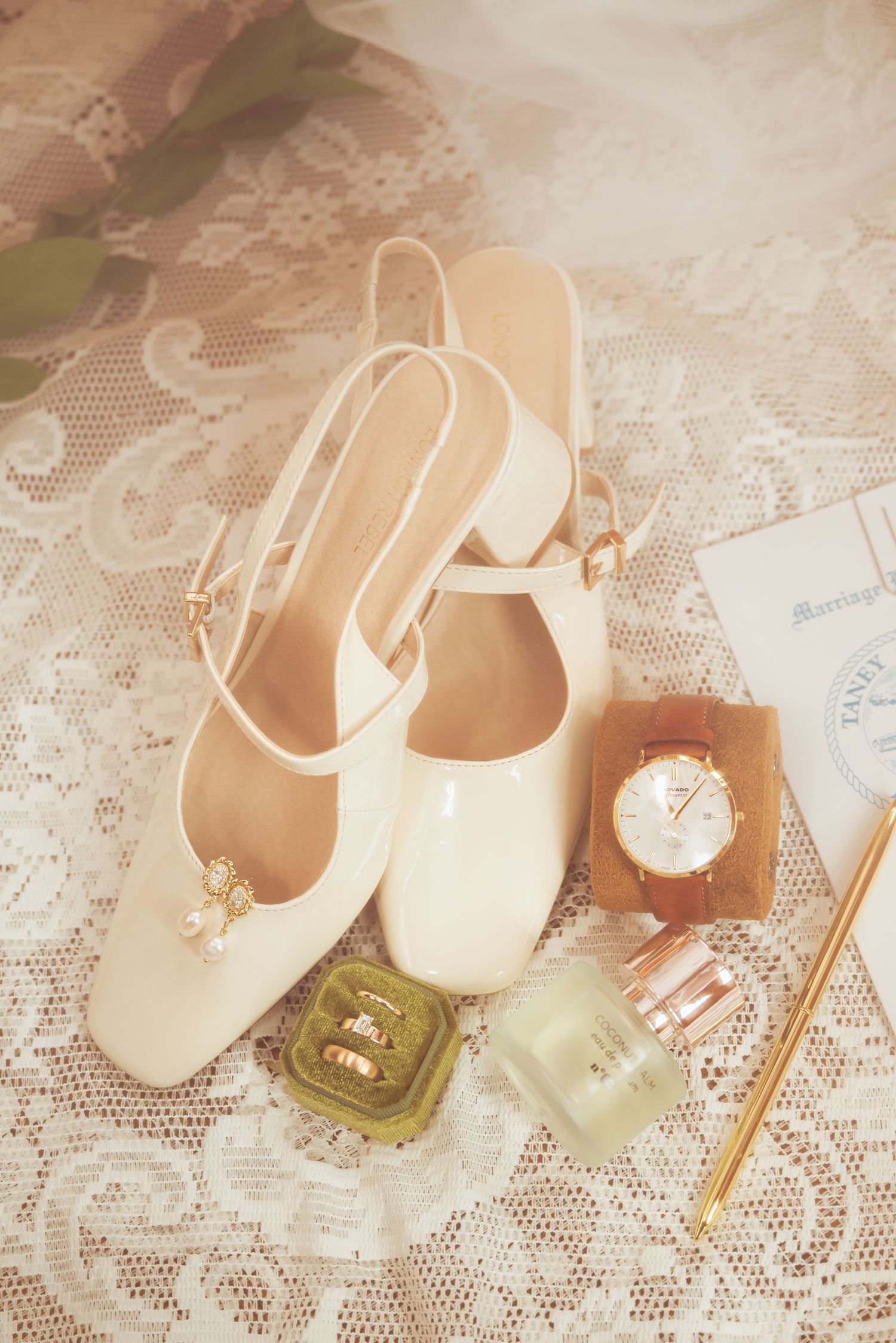 Flat lay of white bridal heels, green velvet ring box, perfume, and a gold watch on lace fabric.