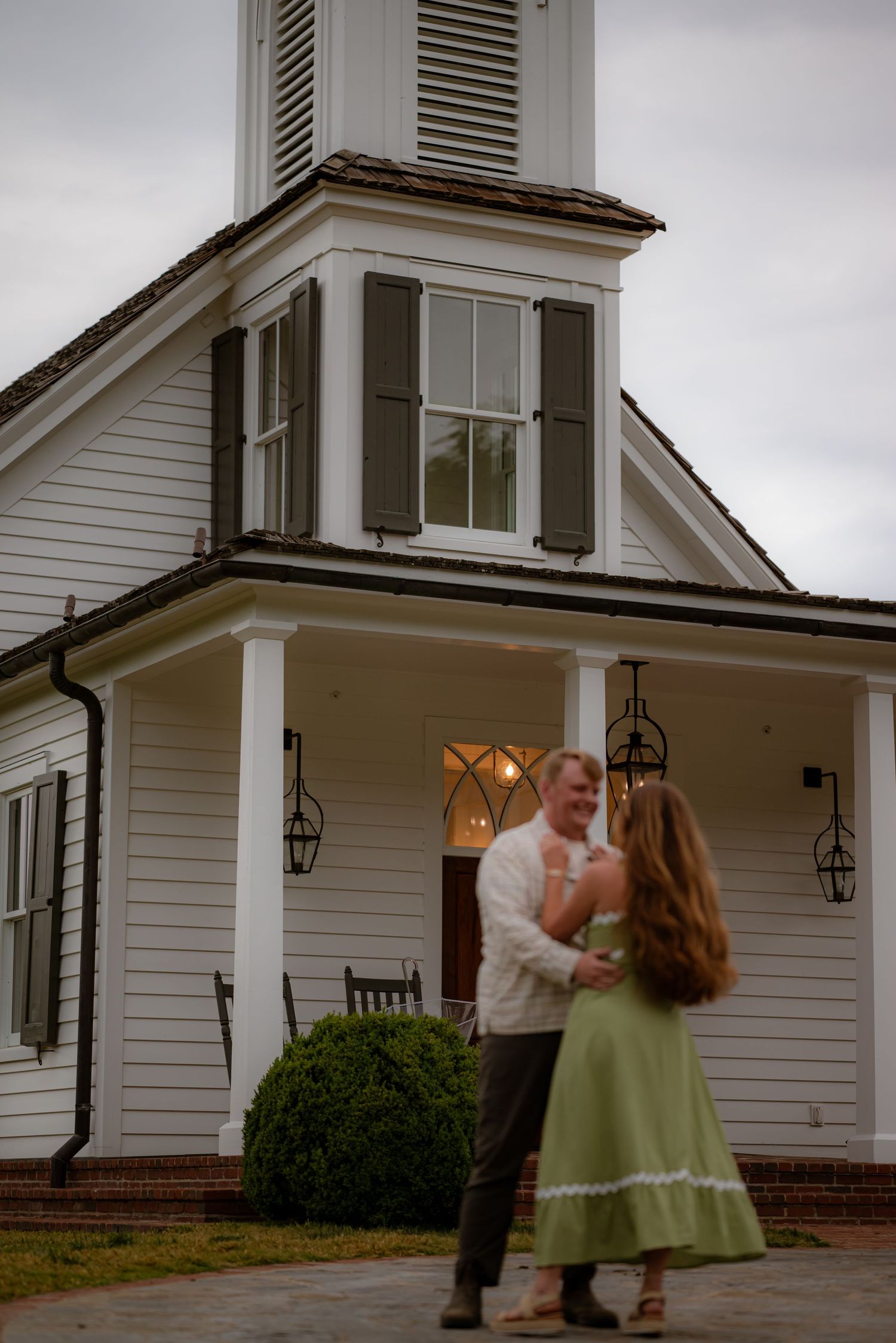 Couple dancing romantically in front of a white colonial church with green shutters at dusk.