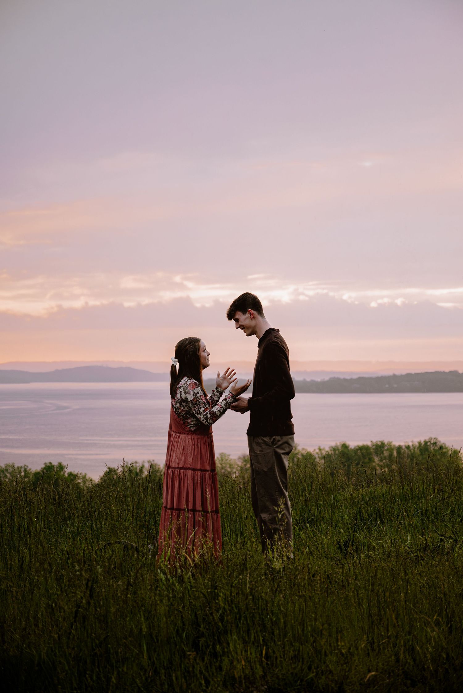 Newly engaged couple standing face to face holding hands in a grassy field with a pastel sunset sky.