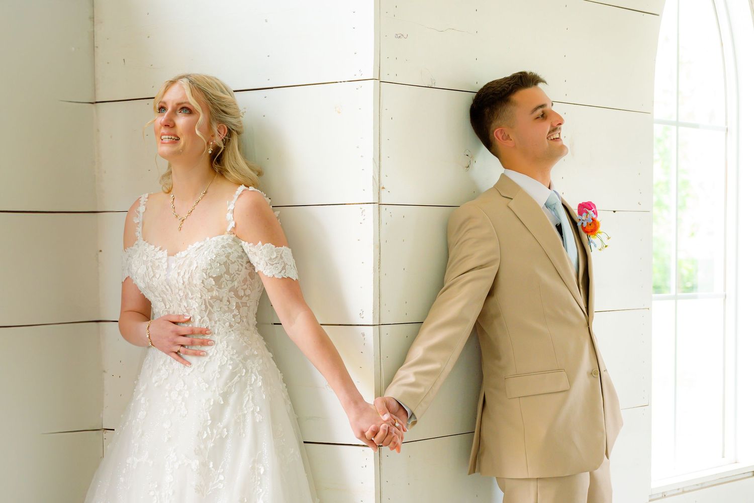 Bride in white lace gown and groom in tan suit hold hands back-to-back against white wall columns.