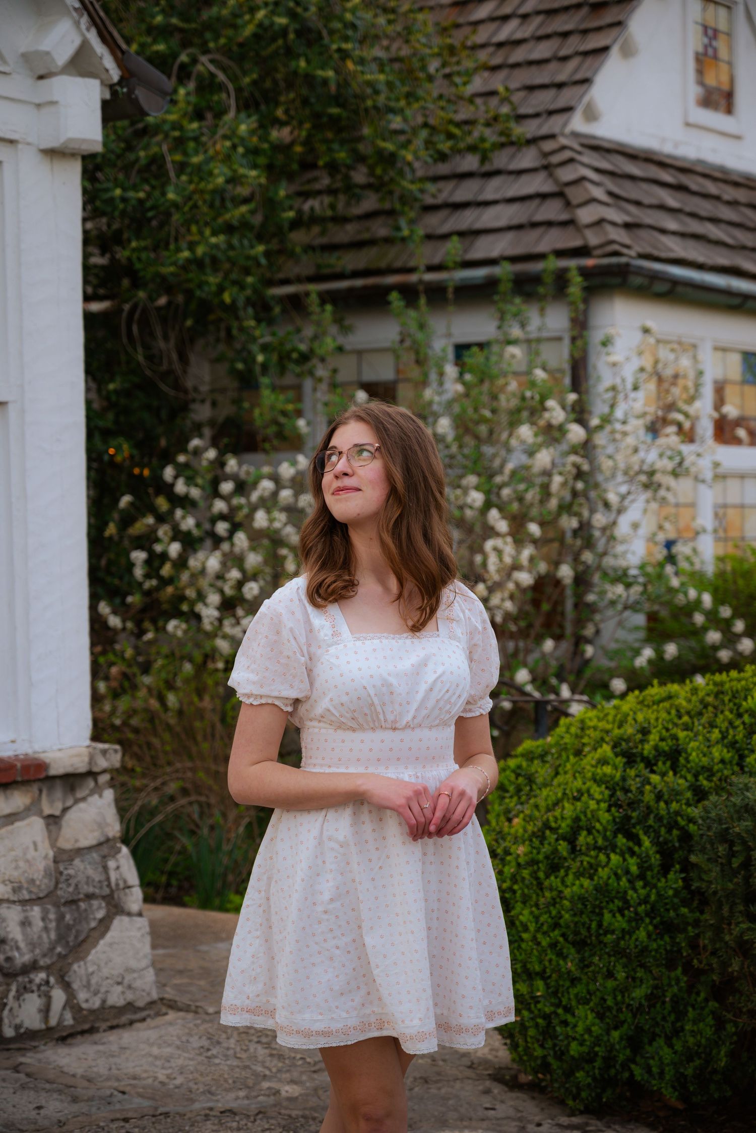 Young woman in a white floral mini dress stands in a charming garden with blooming flowers.