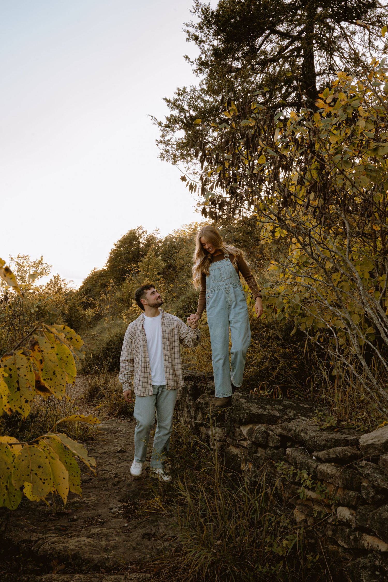 Couple holding hands on a rustic stone path surrounded by autumn foliage during golden hour.
