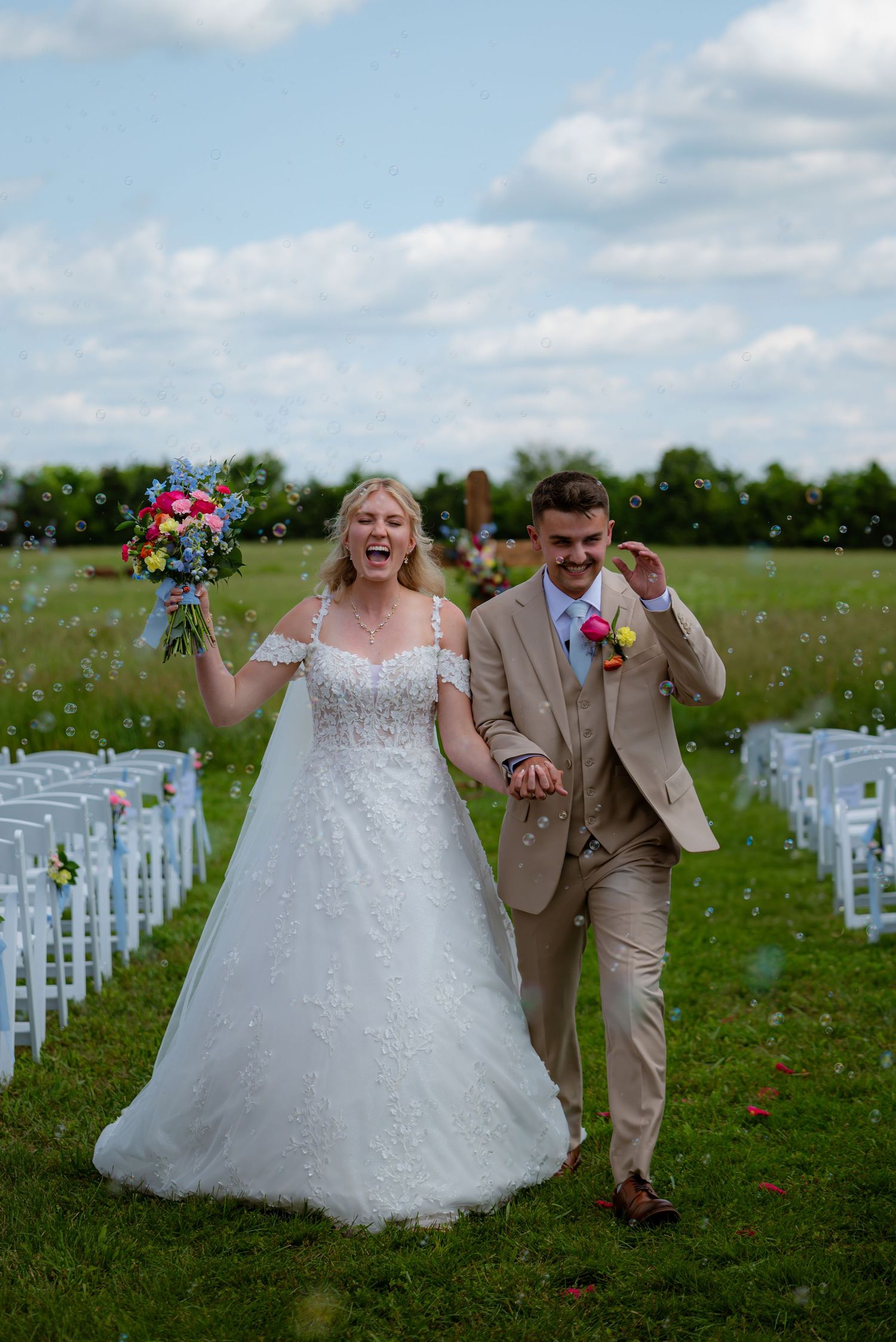Elated newlyweds walking through bubbles down outdoor aisle after sunny field wedding ceremony.