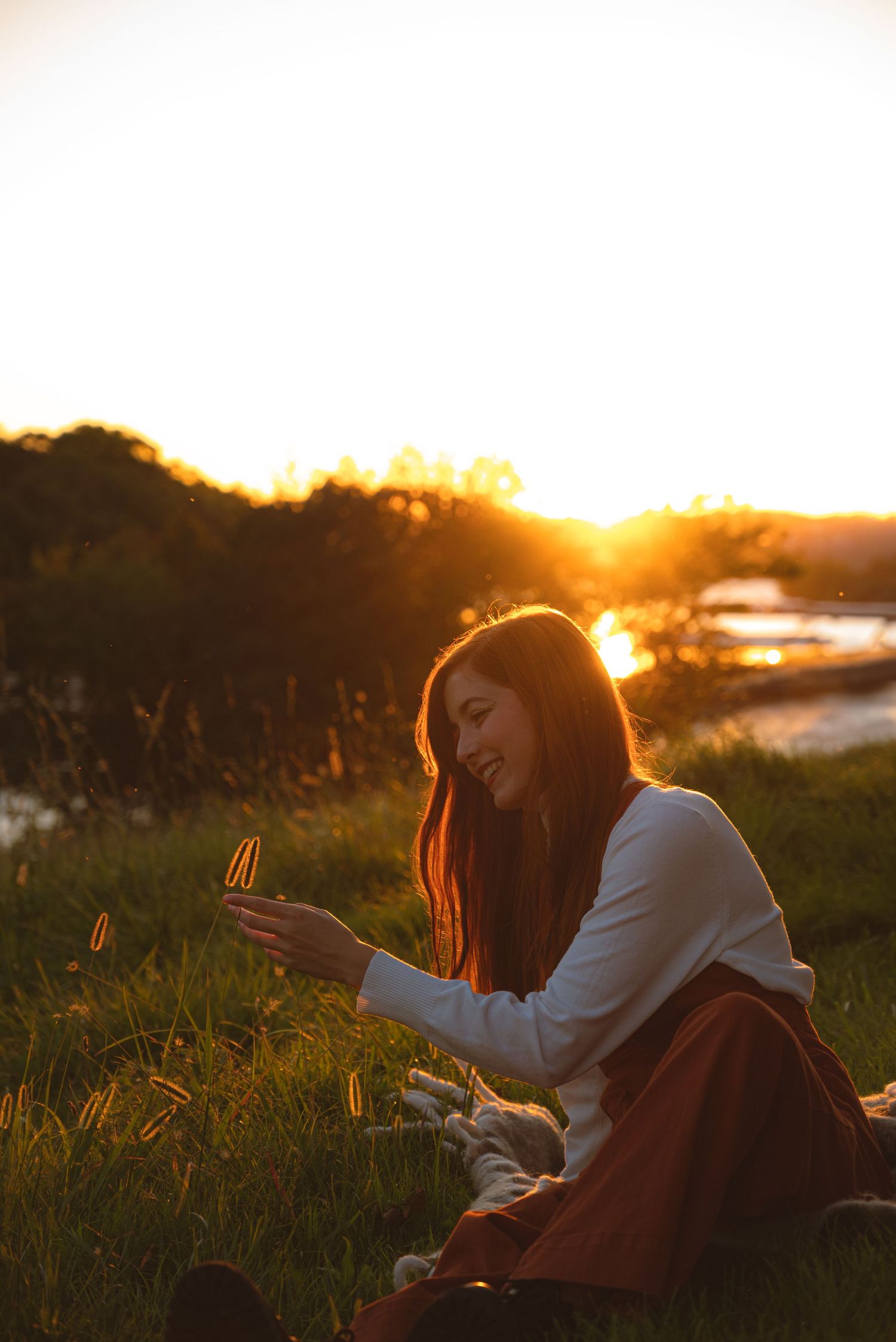 Young woman in white sweater and rust skirt smiles while sitting in grass at golden sunset.