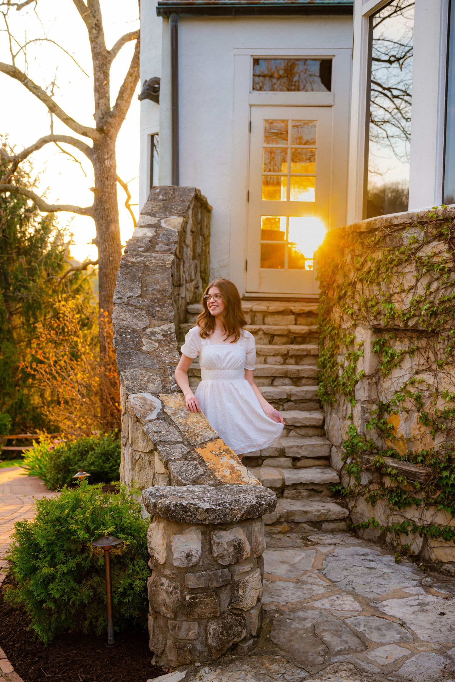 Young woman in white dress smiles on stone steps outside a sunlit cottage at golden hour.