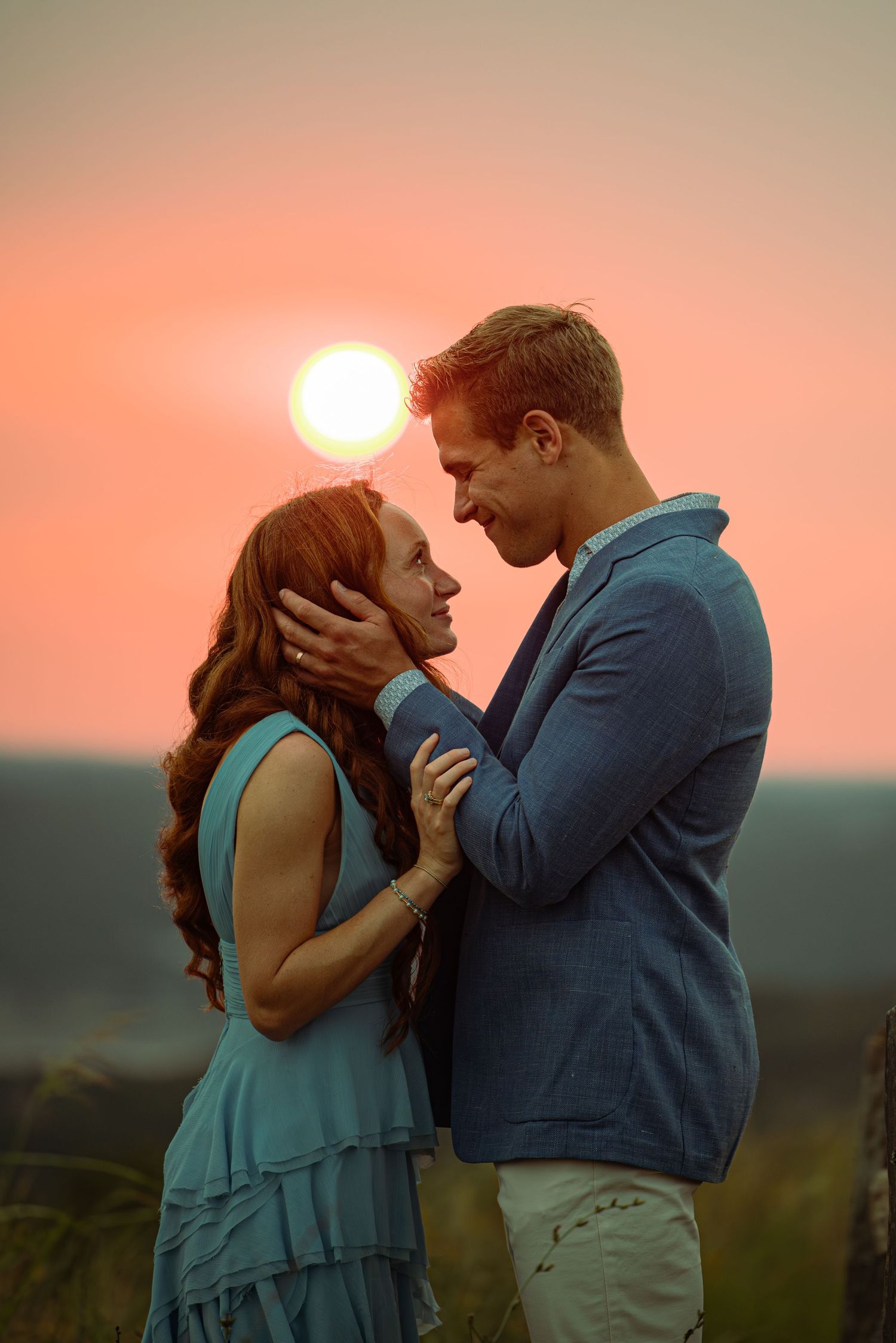 Romantic couple embracing at sunset, woman with red hair in teal dress, man in blue shirt, golden sky background.