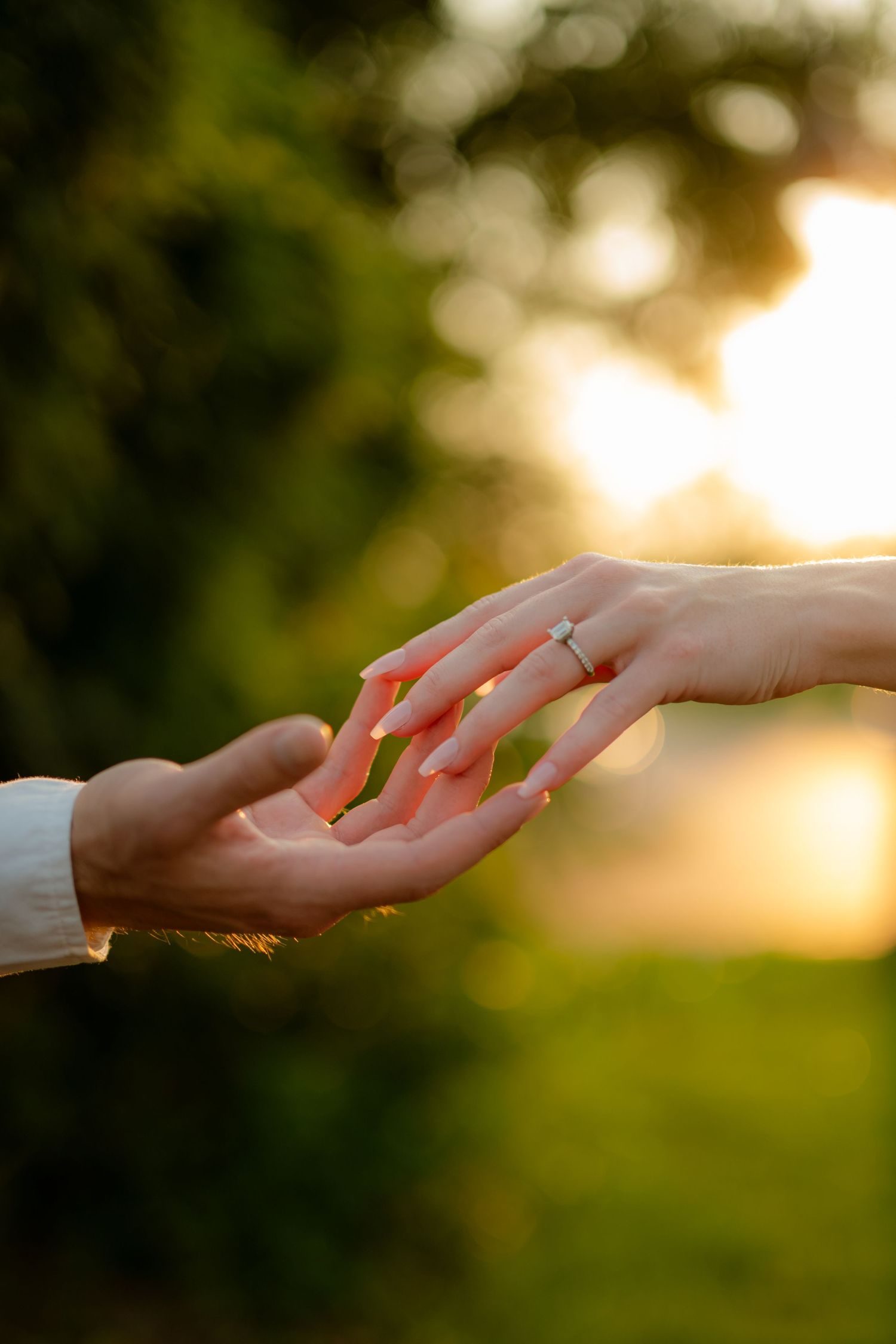 Two hands touching at sunset, woman wearing a diamond engagement ring, romantic proposal moment outdoors.