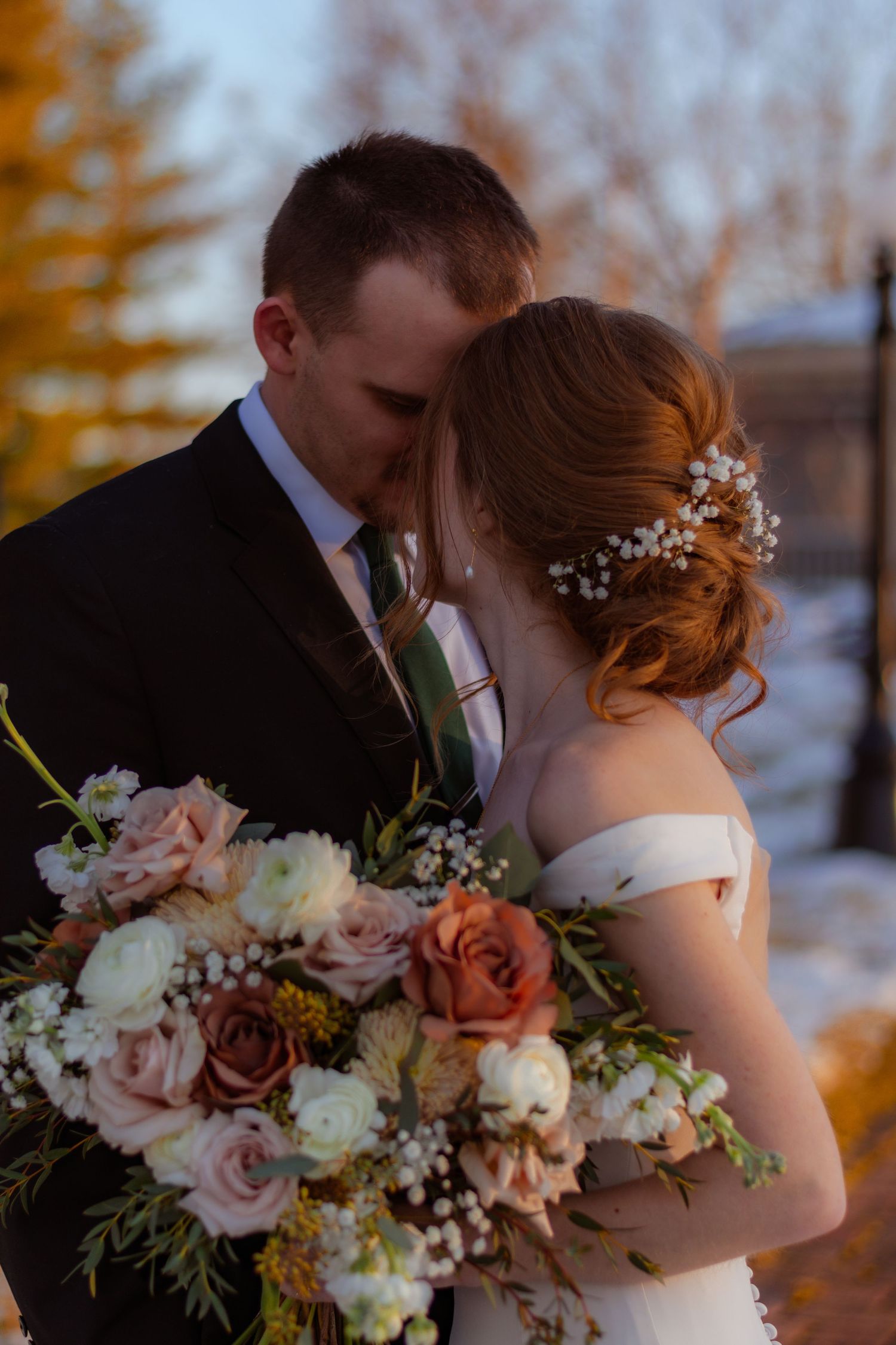 Groom tenderly kisses bride with floral hair piece holding blush and peach bouquet at sunset.