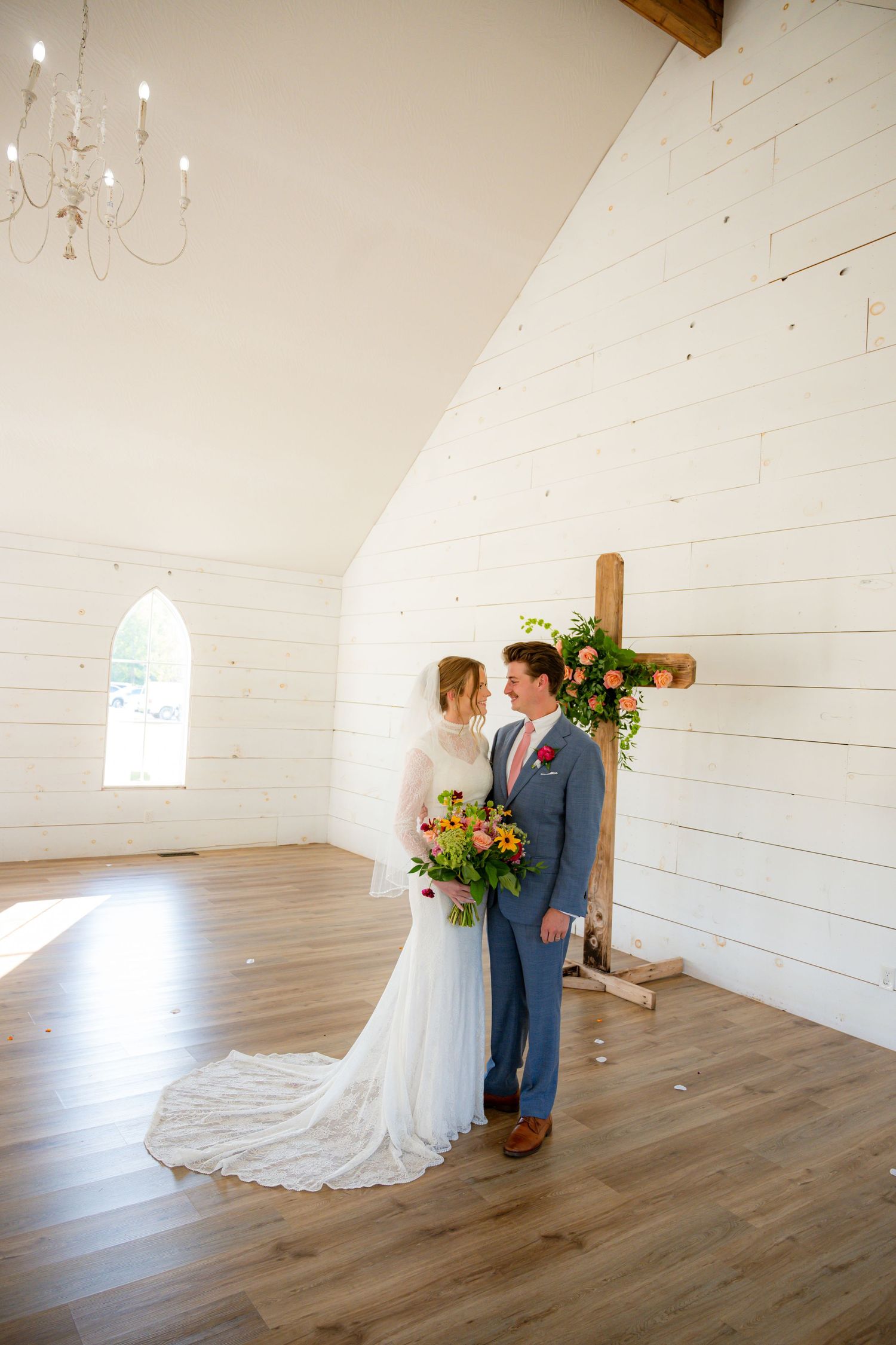 Bride and groom standing together in a white chapel with a wooden cross adorned with flowers and a chandelier overhead.
