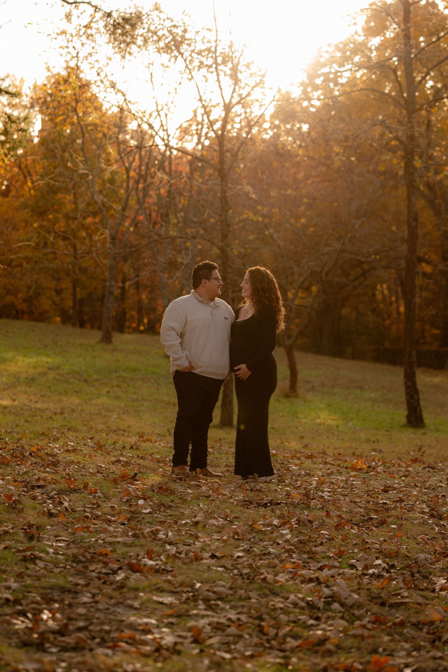 Couple stands facing each other in autumn park at golden hour, surrounded by fallen leaves and colorful trees.
