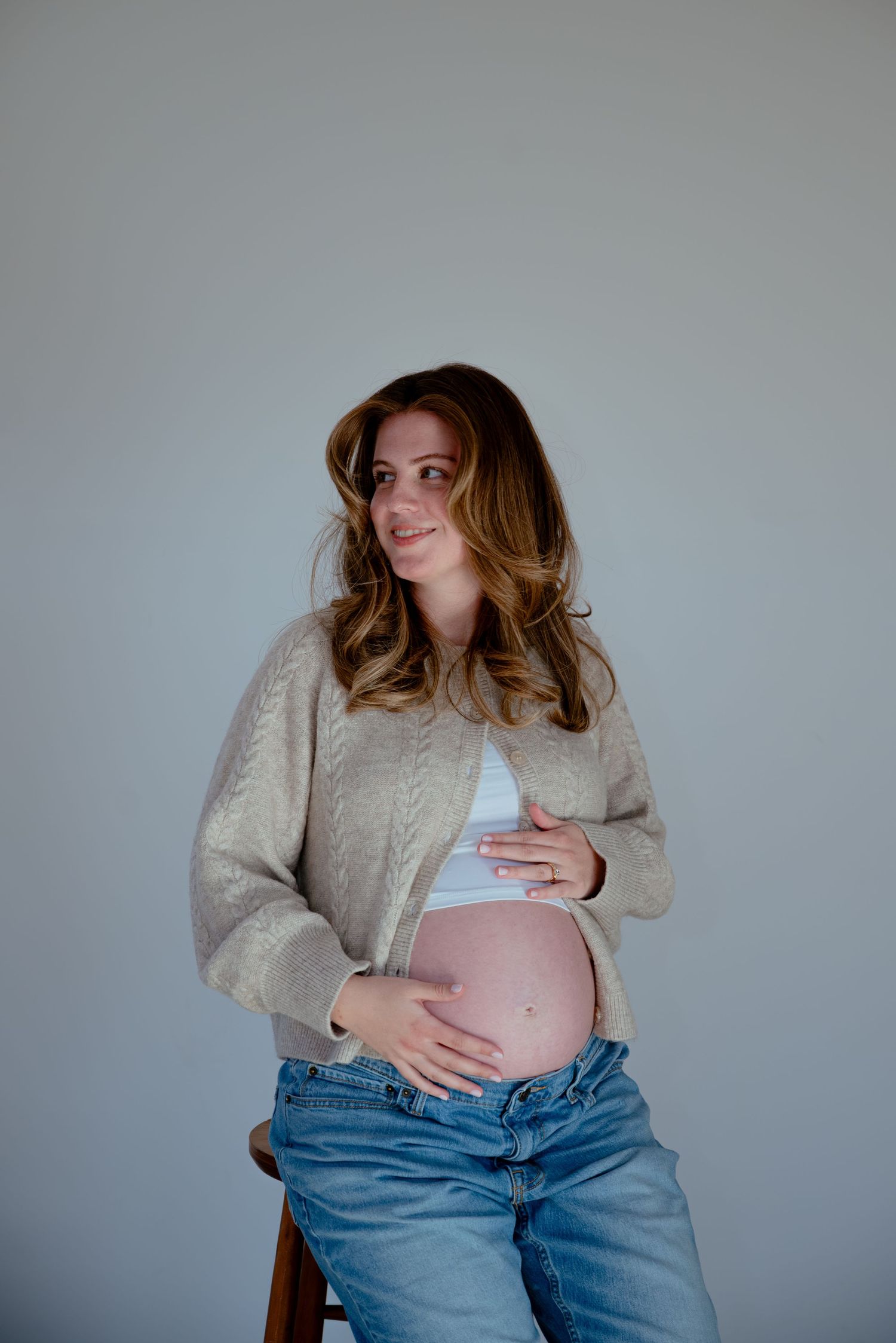 Pregnant woman in beige cardigan and jeans cradling baby bump while sitting on wooden stool.