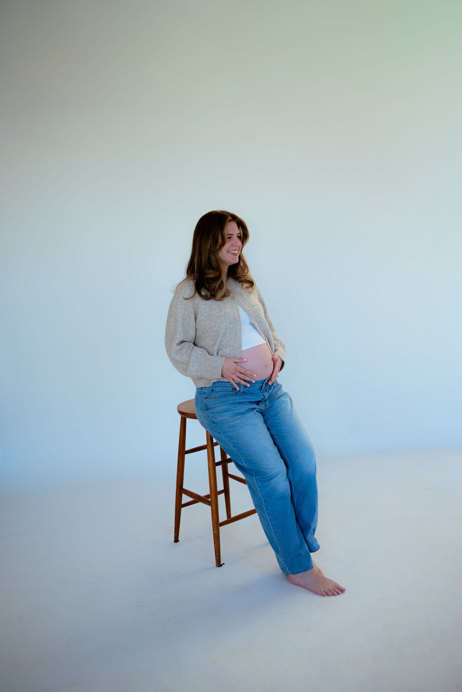 Smiling pregnant woman in beige cardigan and jeans sitting on wooden stool in bright studio.