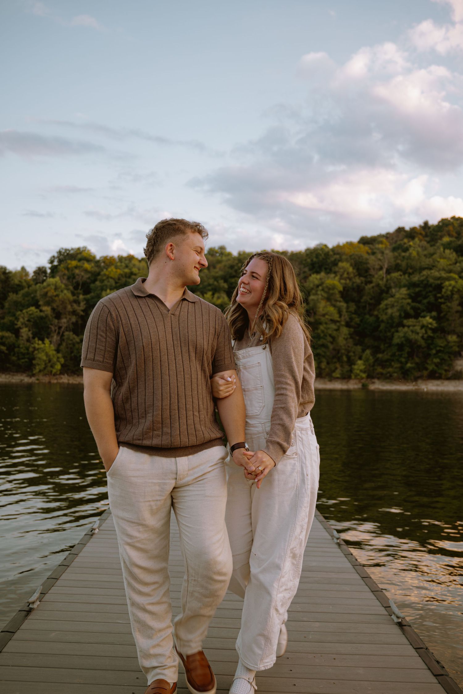 Happy couple walking hand-in-hand on a lakeside dock at dusk, smiling at each other.