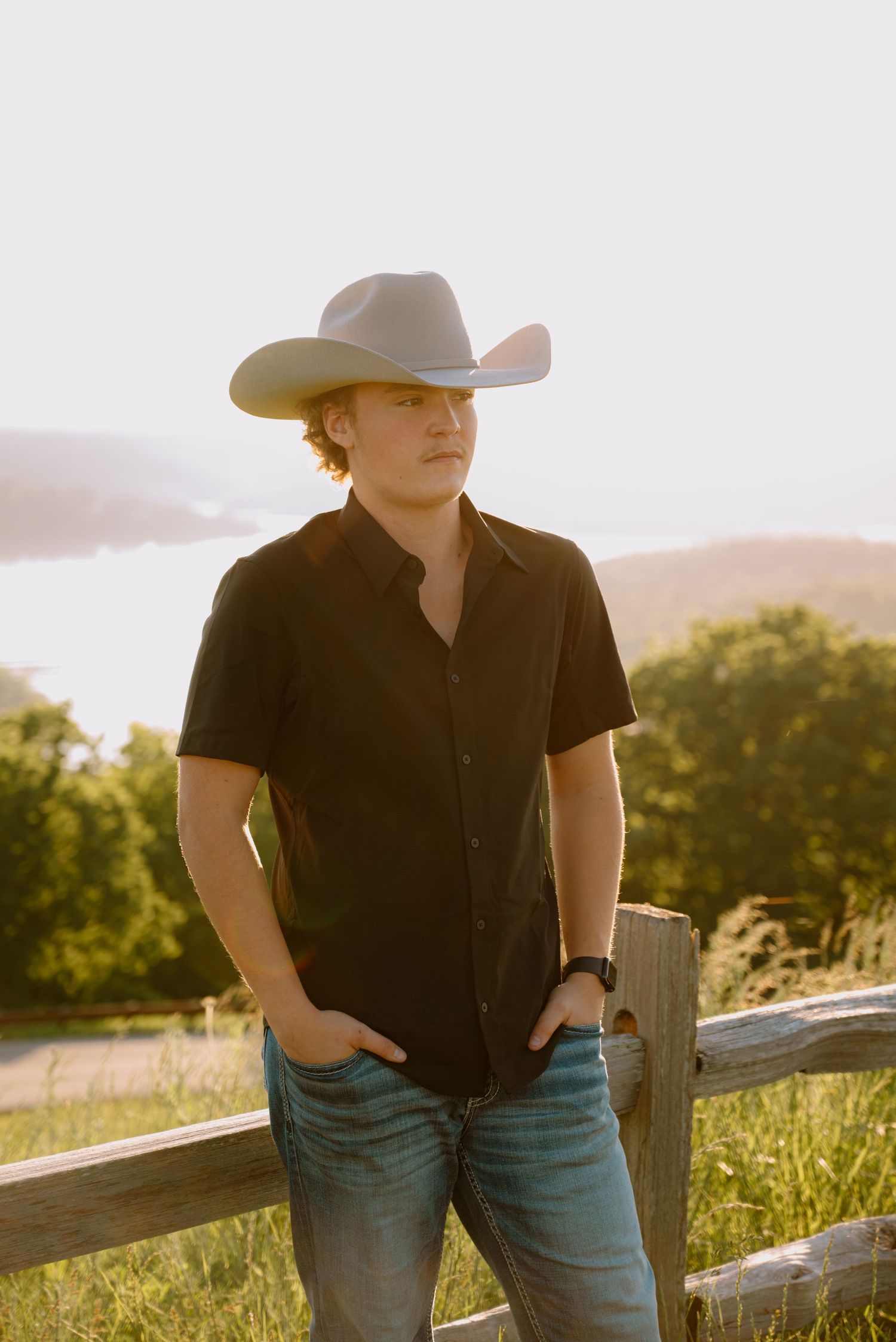A young man wearing a white cowboy hat and black shirt stands by a wooden fence at golden hour.