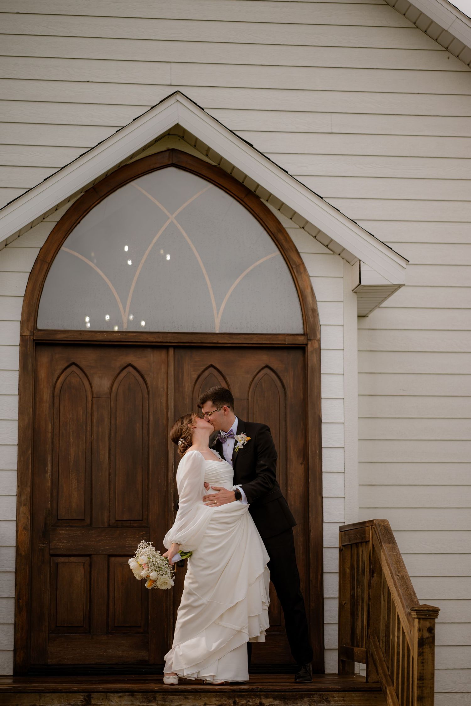 Bride and groom share a kiss in front of elegant wooden chapel doors on their wedding day.