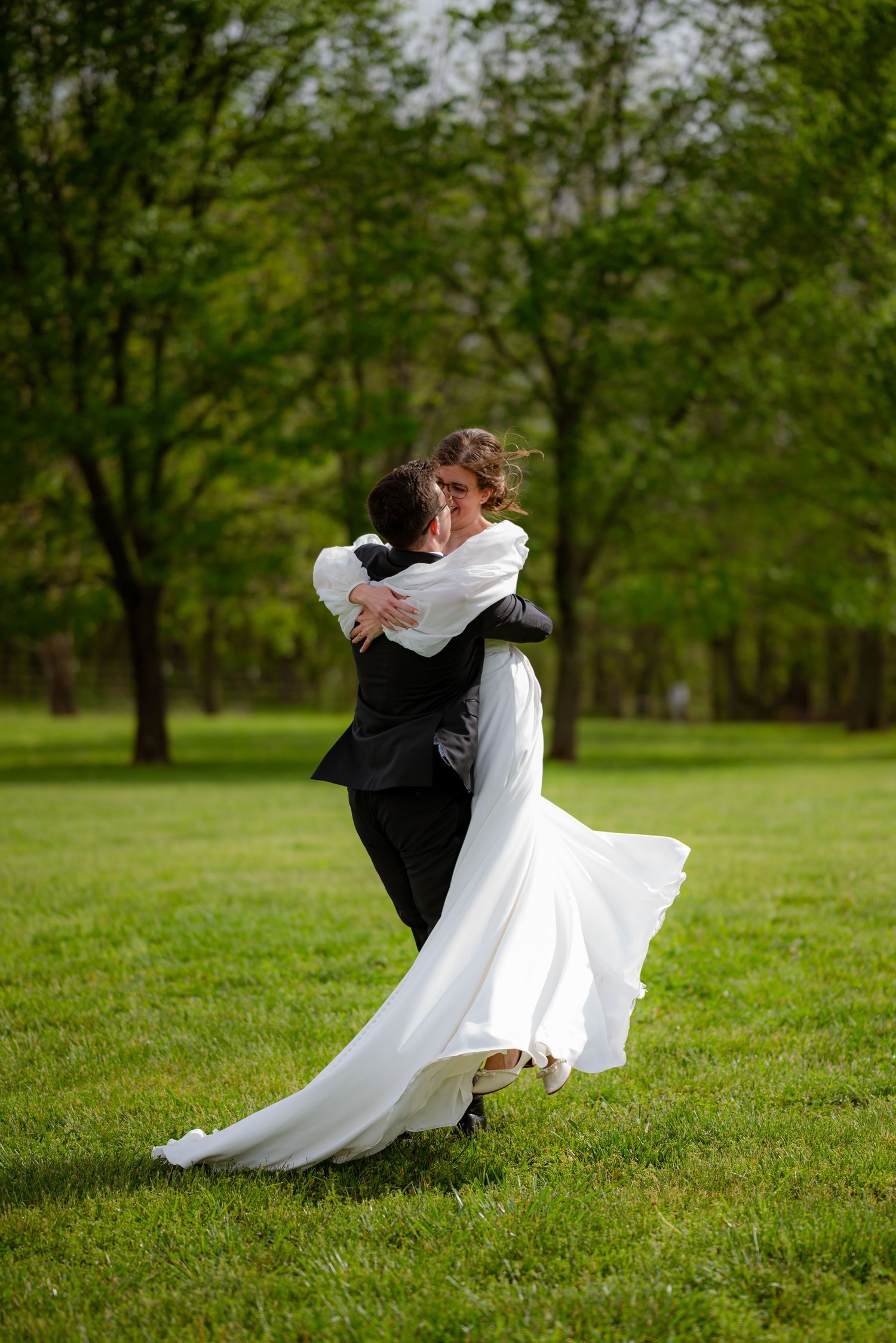 Bride in white gown being lifted by groom in black suit, dancing joyfully on green grass with trees in background.