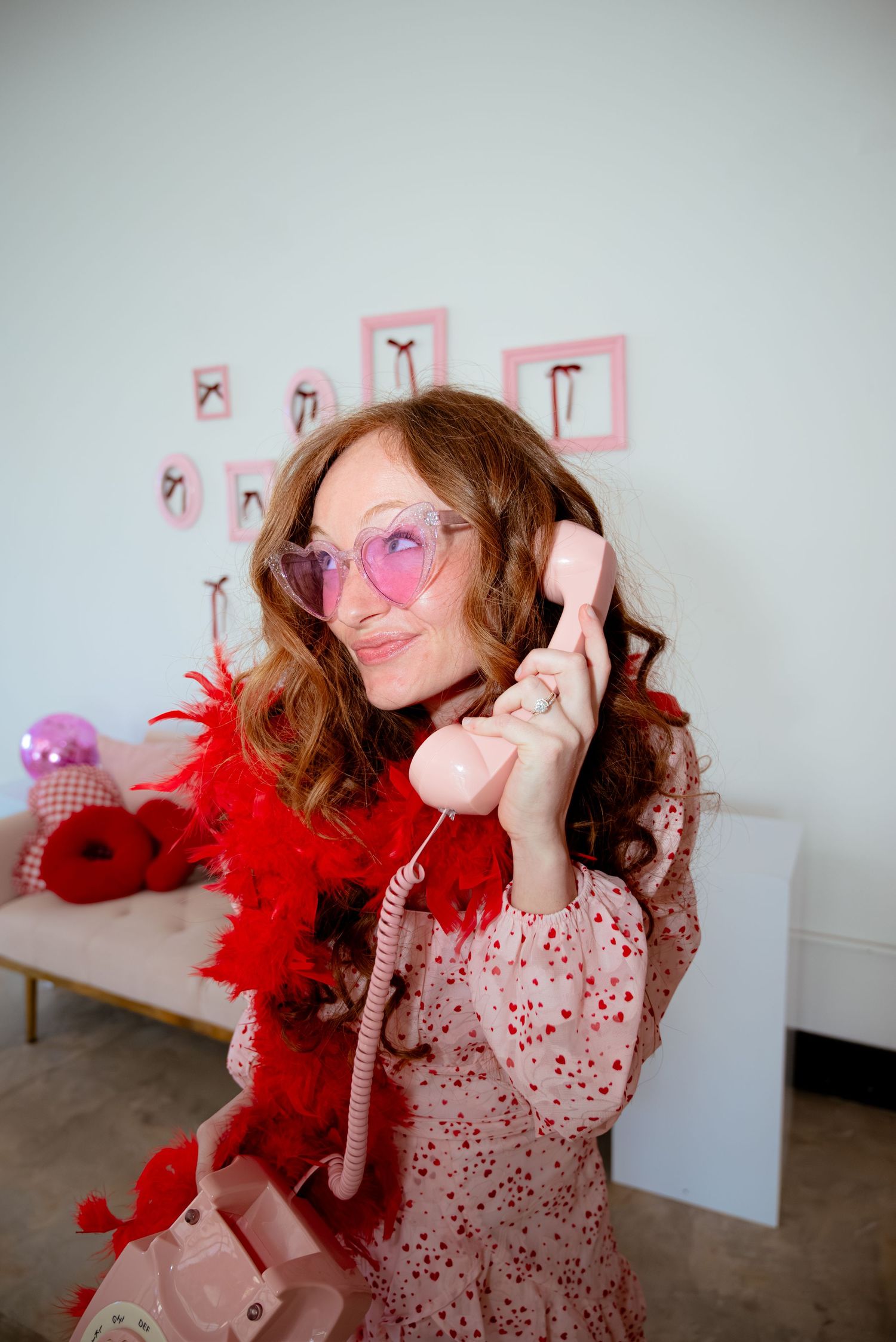 Smiling woman in pink heart sunglasses and red feather boa holding a pink telephone receiver.