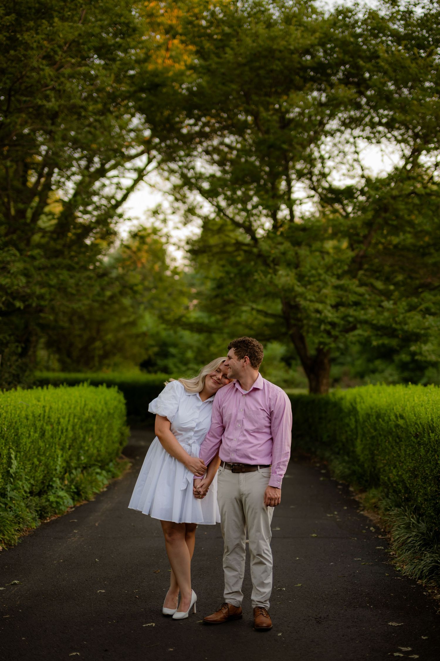 A couple shares a tender moment on a tree-lined path, woman in white dress, man in pink shirt holding hands.