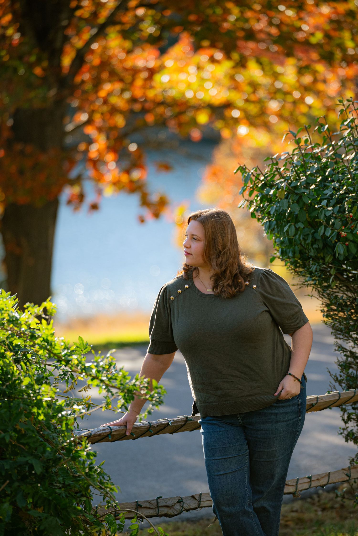 Woman in olive top leaning on rustic wooden fence surrounded by vibrant autumn foliage near a lake.