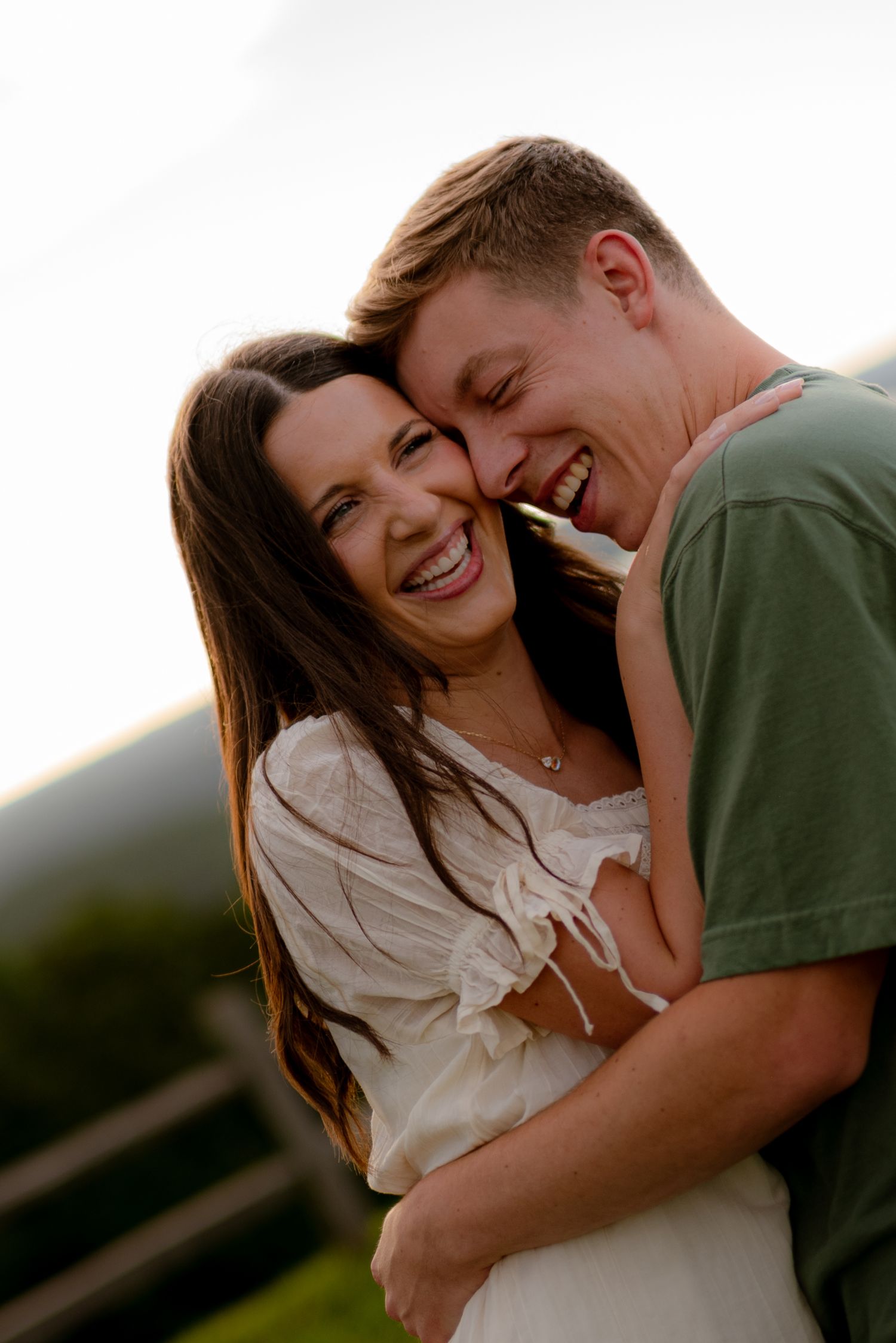 A joyful couple laughs and embraces outdoors, the woman in white and man in green, with a scenic mountain backdrop.