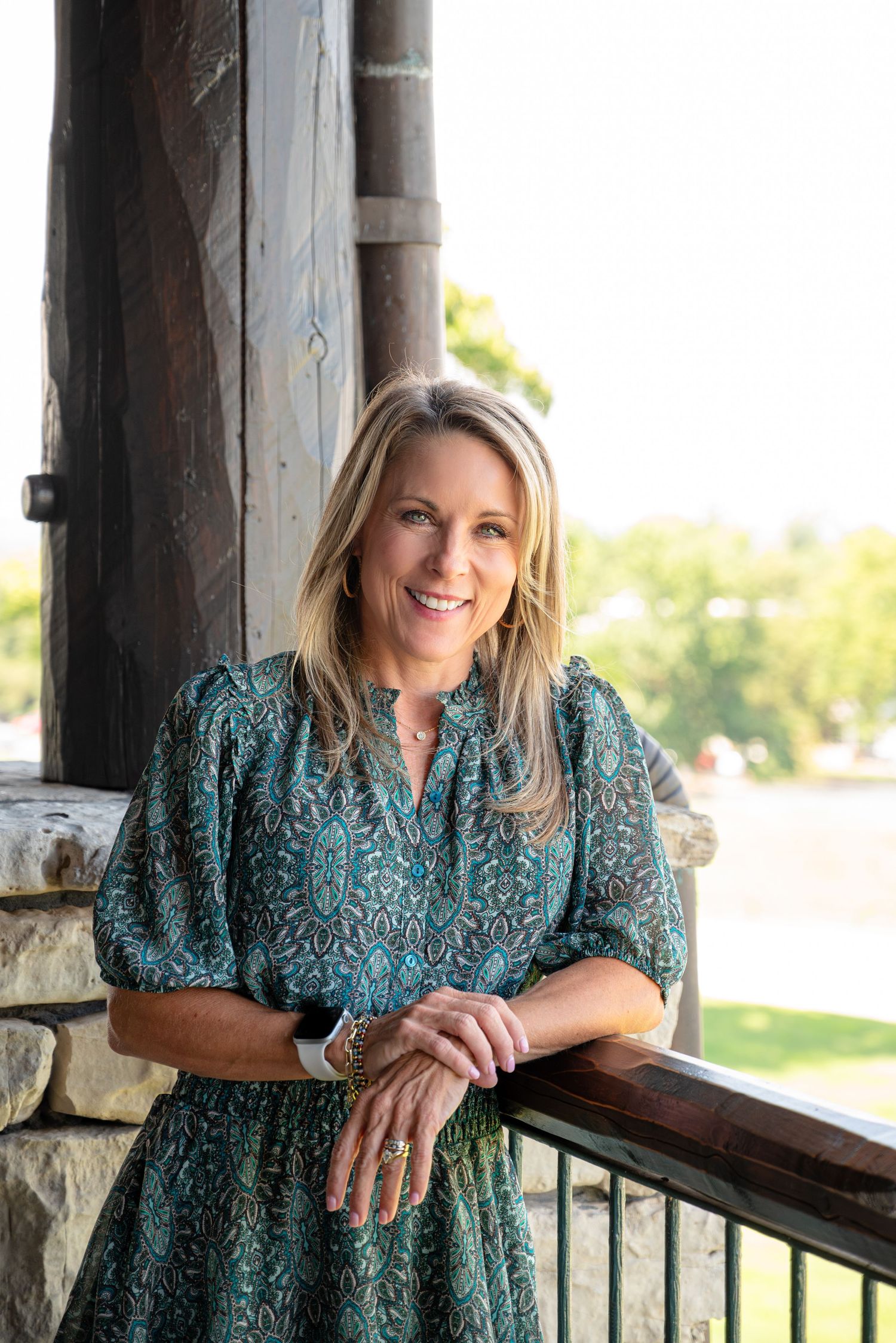 Smiling blonde woman in a teal floral dress leaning on a wooden railing on an outdoor porch.