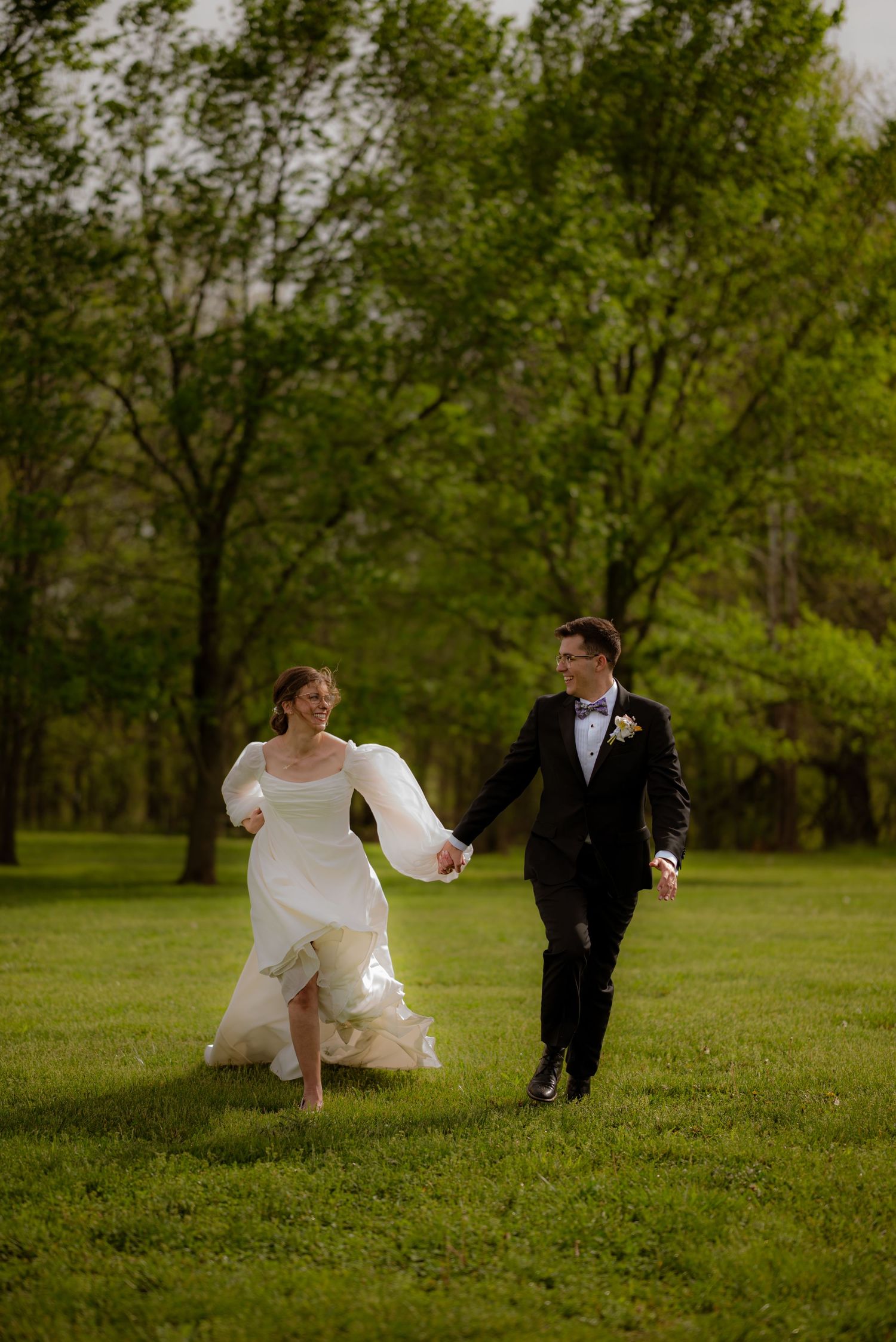 A joyful bride and groom hold hands while running through a lush green park on their wedding day.