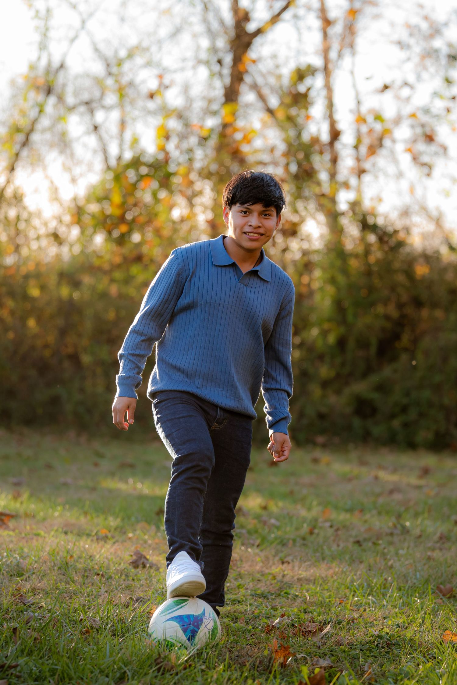 Smiling teen boy in blue polo sweater kicking soccer ball in autumn park setting.