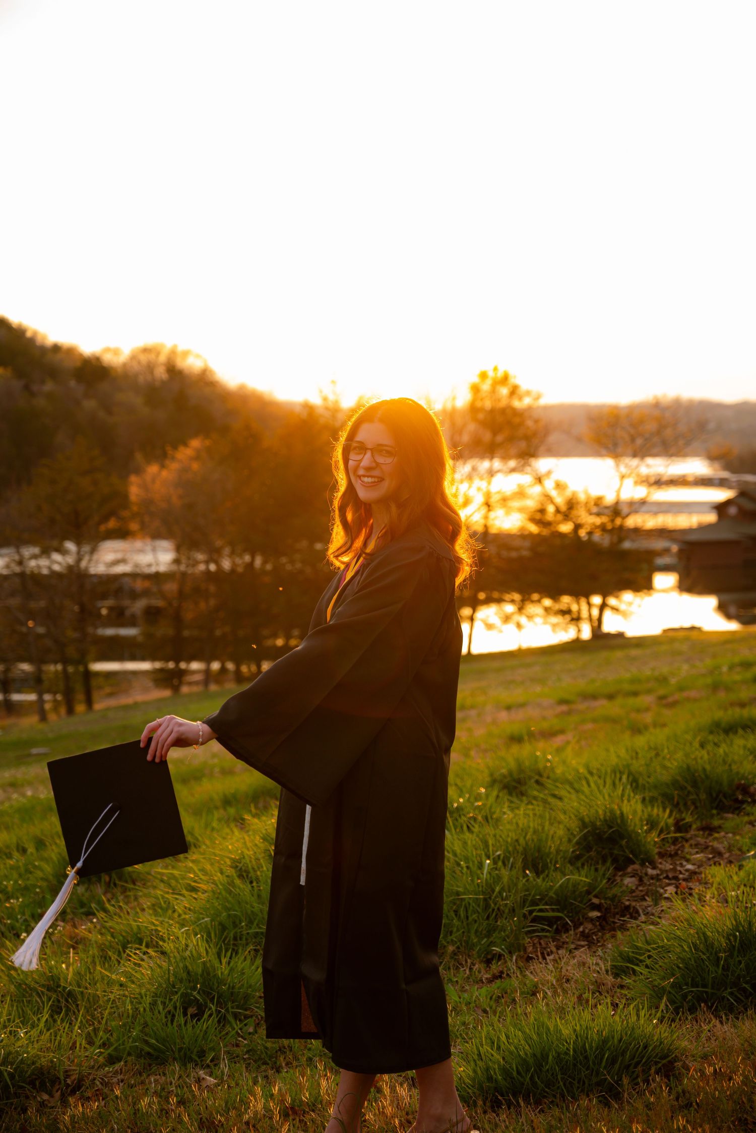 Graduate in black cap and gown holds mortarboard at golden sunset by a peaceful lakeside.