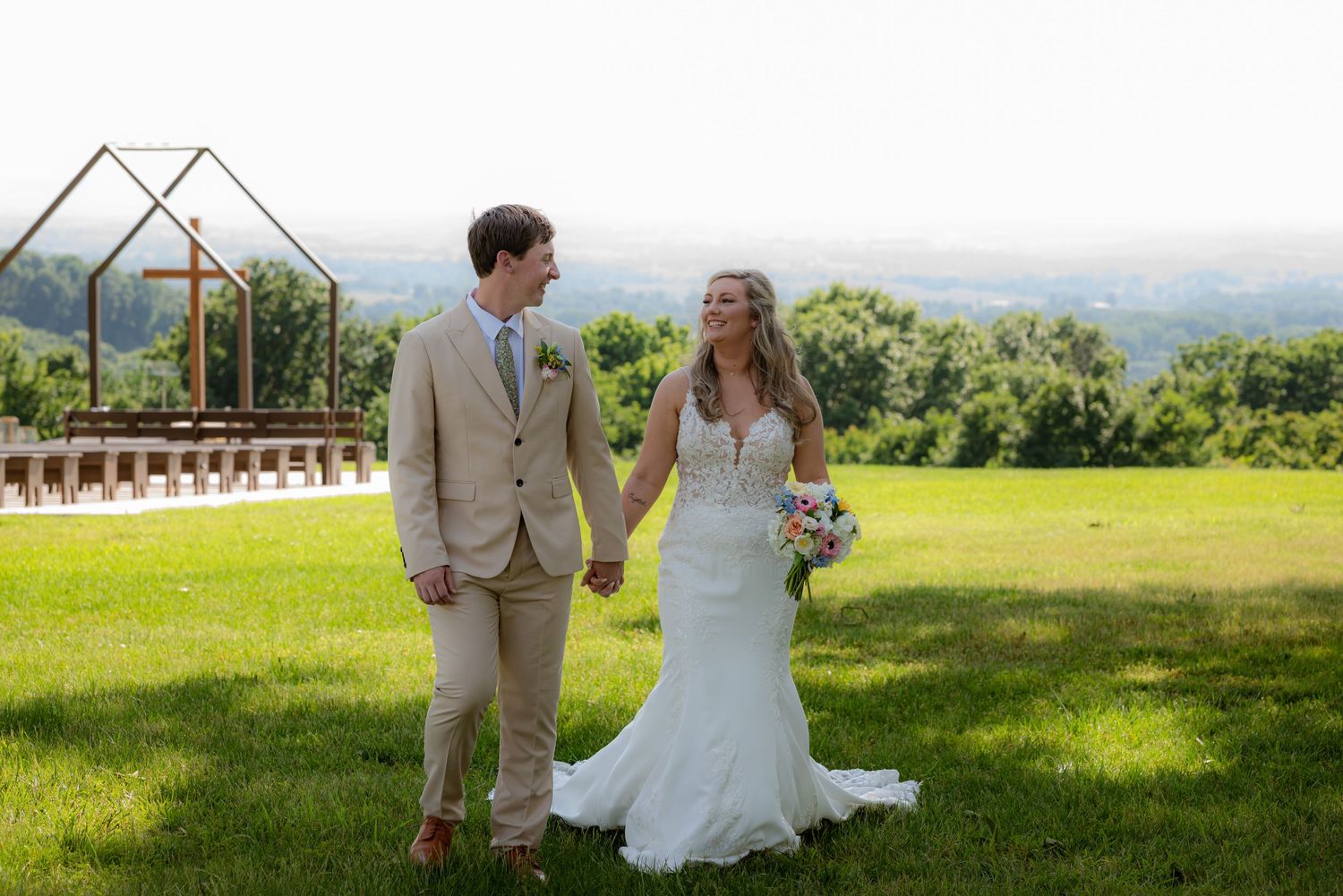 Bride and groom holding hands on green lawn with outdoor wedding ceremony arch and scenic countryside view.