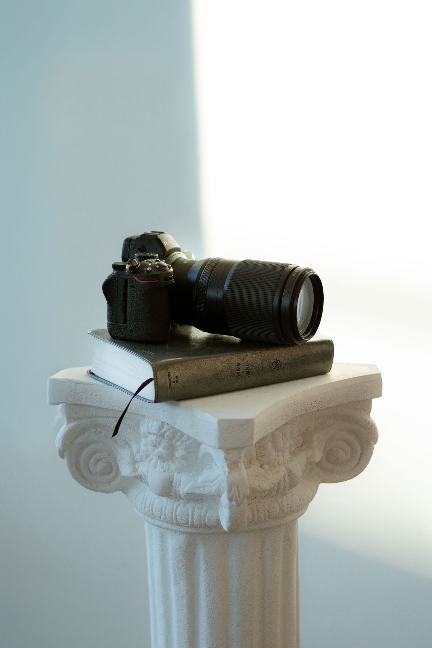 A DSLR camera with telephoto lens resting on books atop a white classical decorative column pedestal.