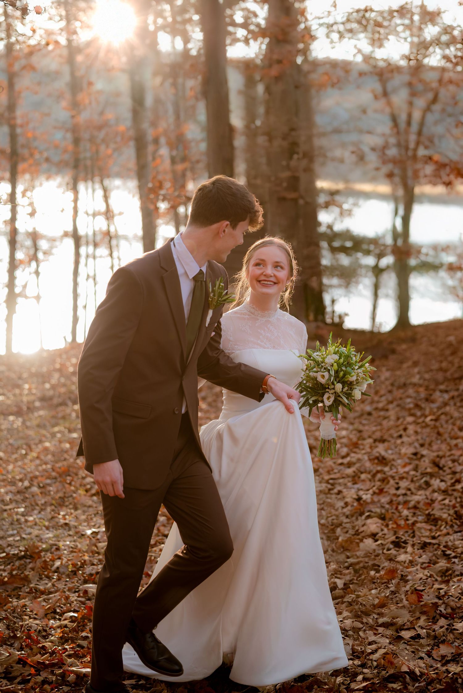 Bride and groom walking through an autumn forest at golden hour near a lake, bride laughing joyfully.