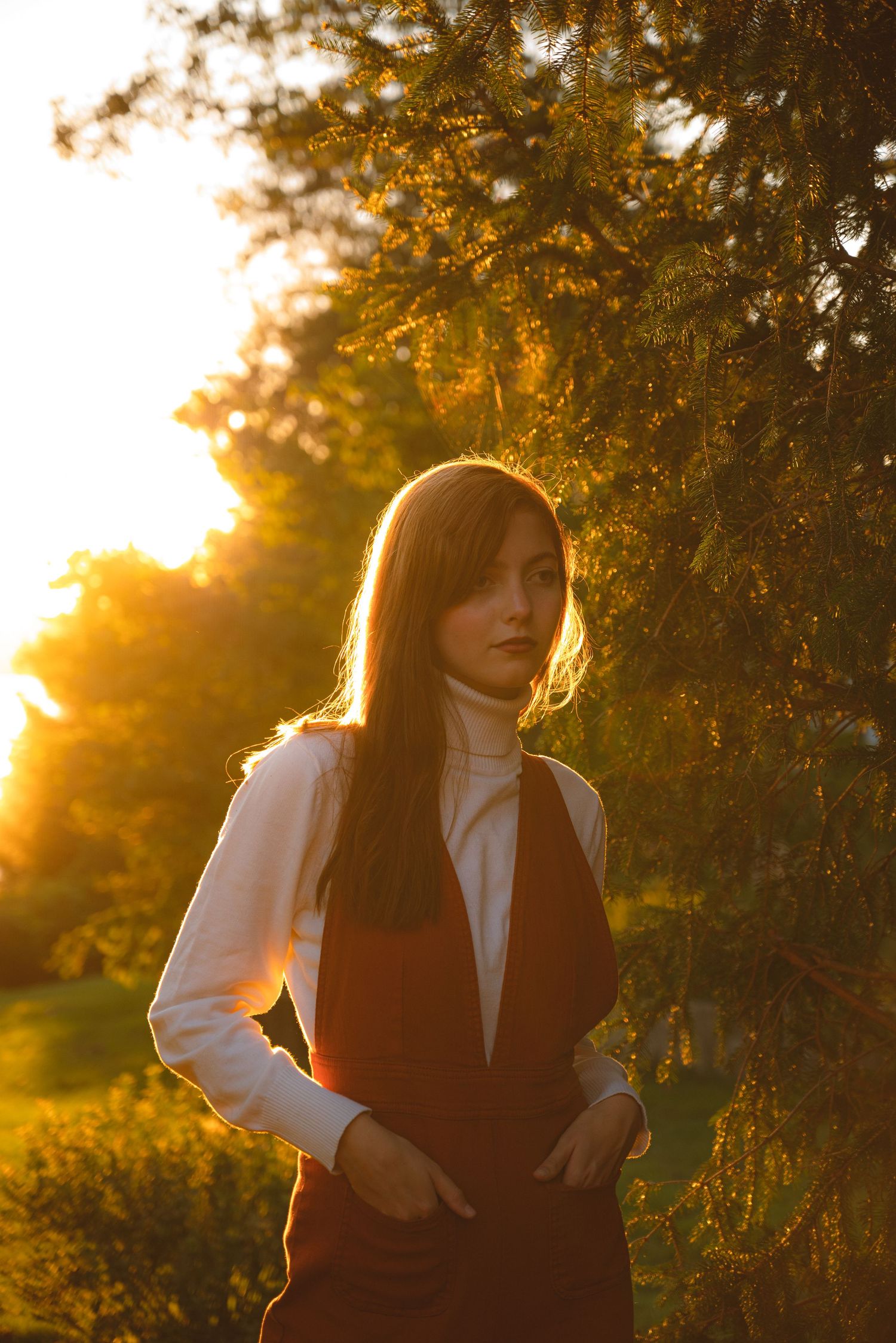 Young woman in rust jumper and white turtleneck stands near evergreen trees at golden sunset.