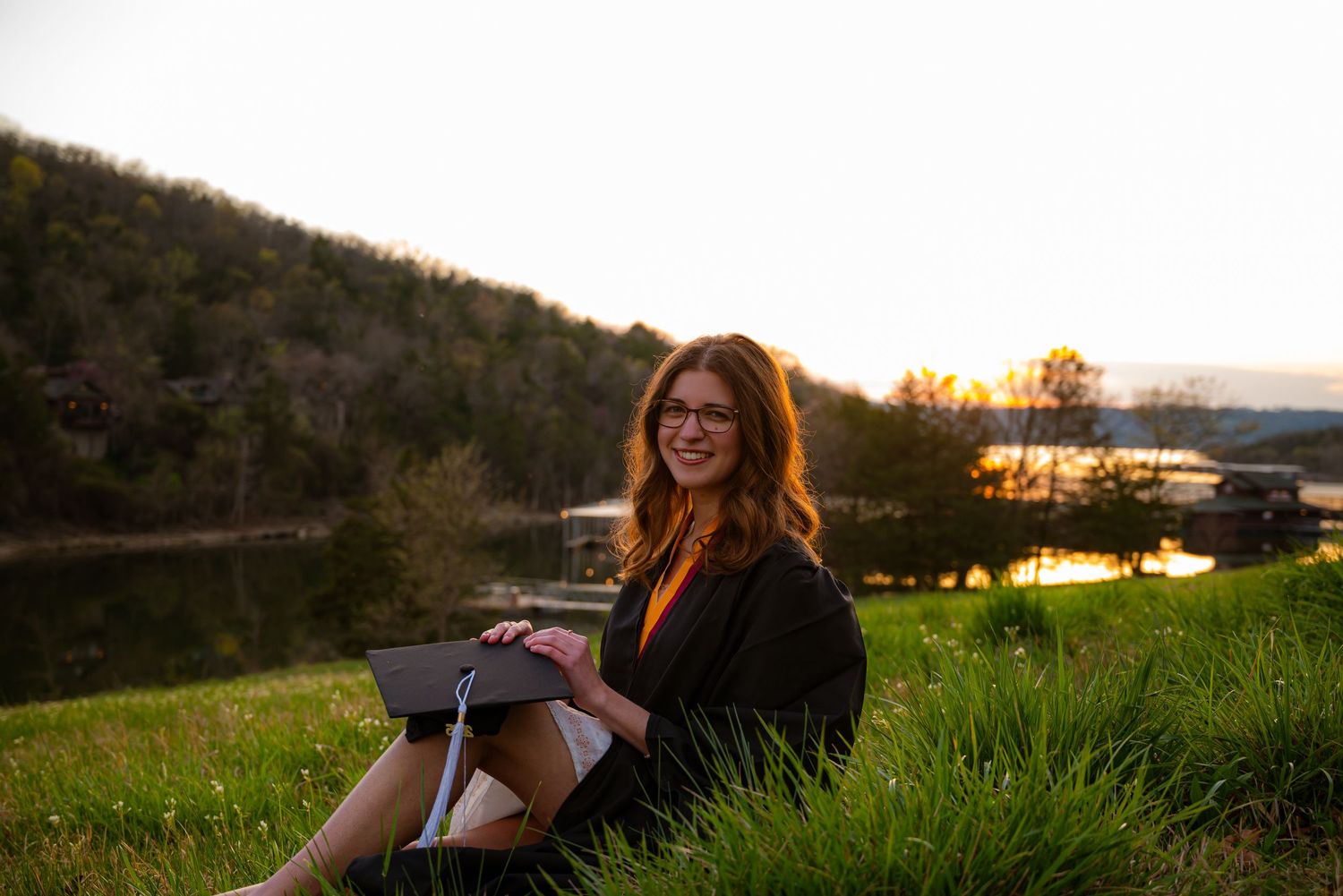 Smiling graduate in black gown sits on grass holding diploma at a beautiful lakeside sunset.