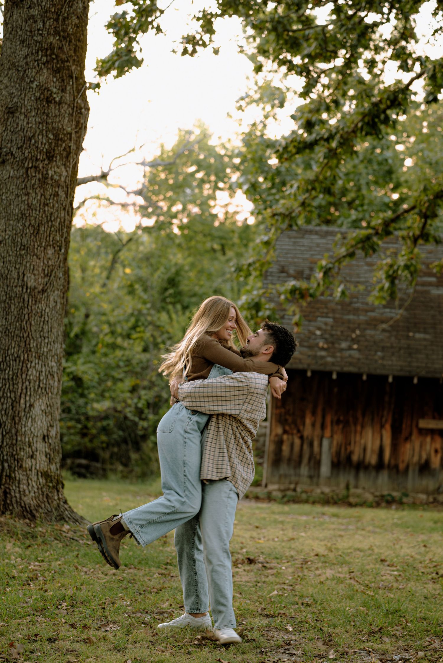 Man lifting woman near large oak tree beside rustic barn in golden hour light.