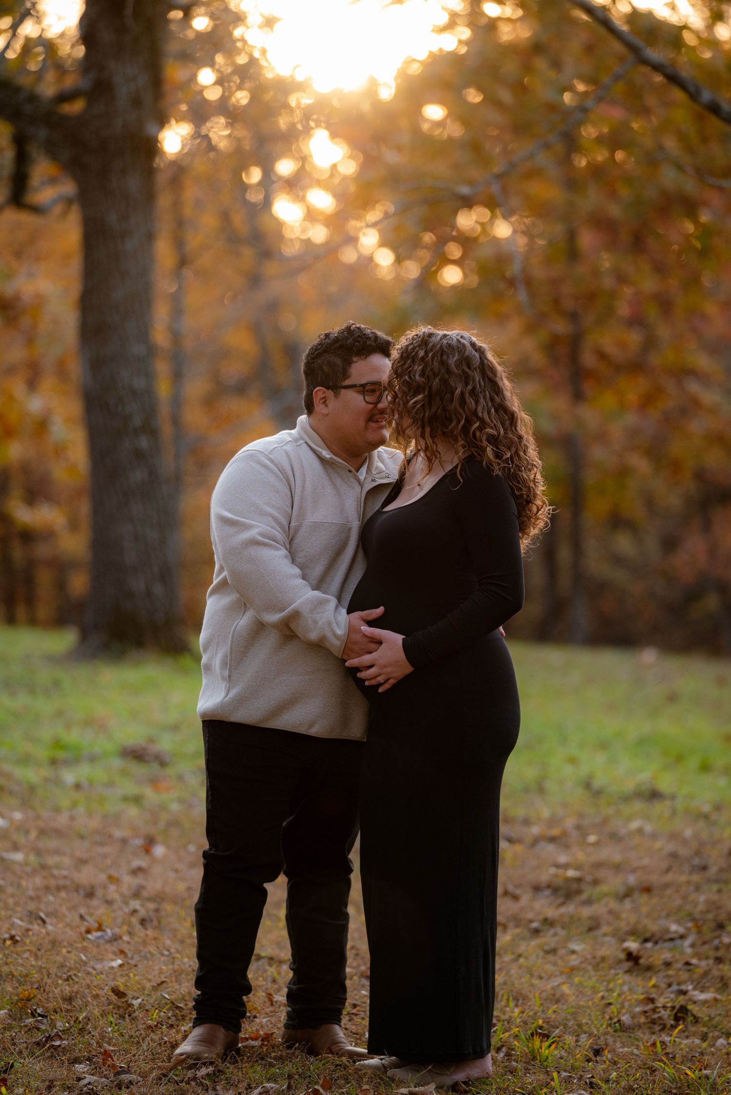 Expecting couple sharing a kiss while cradling baby bump in autumn sunset woods.