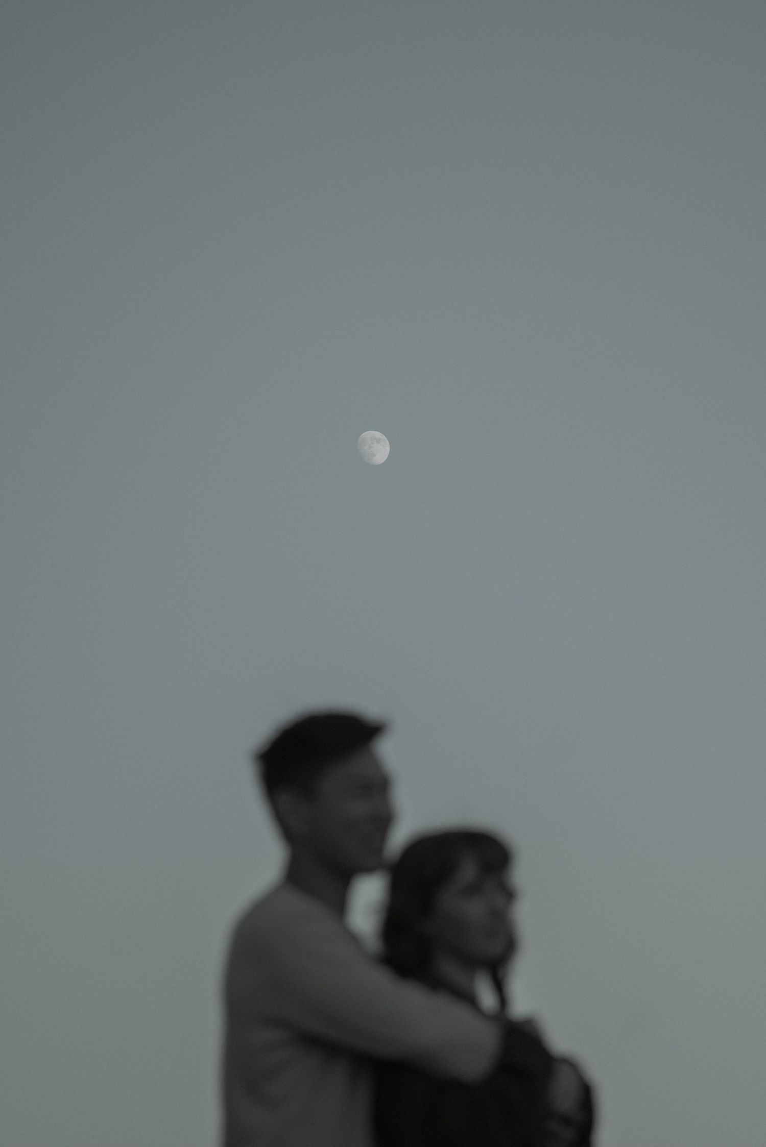 Couple embracing under a glowing moon in a soft monochromatic twilight sky.